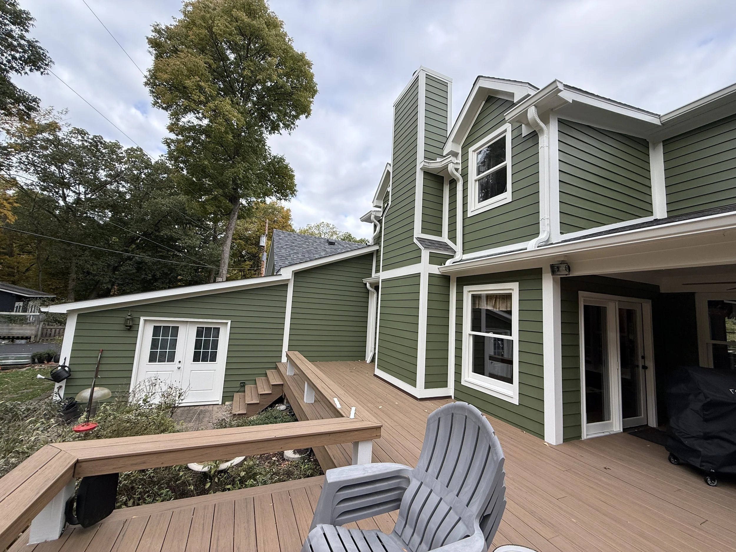 A multi-story green house with white trim and multiple windows, a wooden deck with chairs, and a backyard with trees.