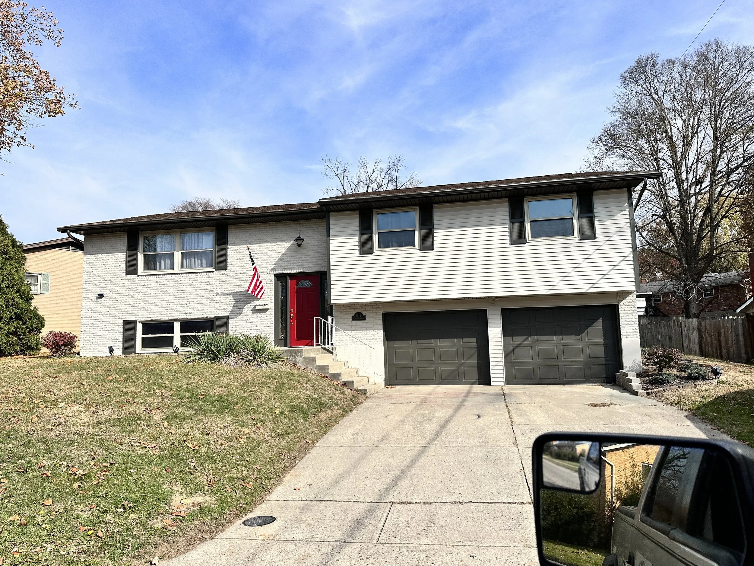 A two-story house with white brick and siding exterior, black shutters, and a red front door. There are two garage doors and a small yard with grass and plants. An American flag is mounted next to the front door. The driveway extends to the street, and the house is surrounded by a wooden fence with trees in the background.