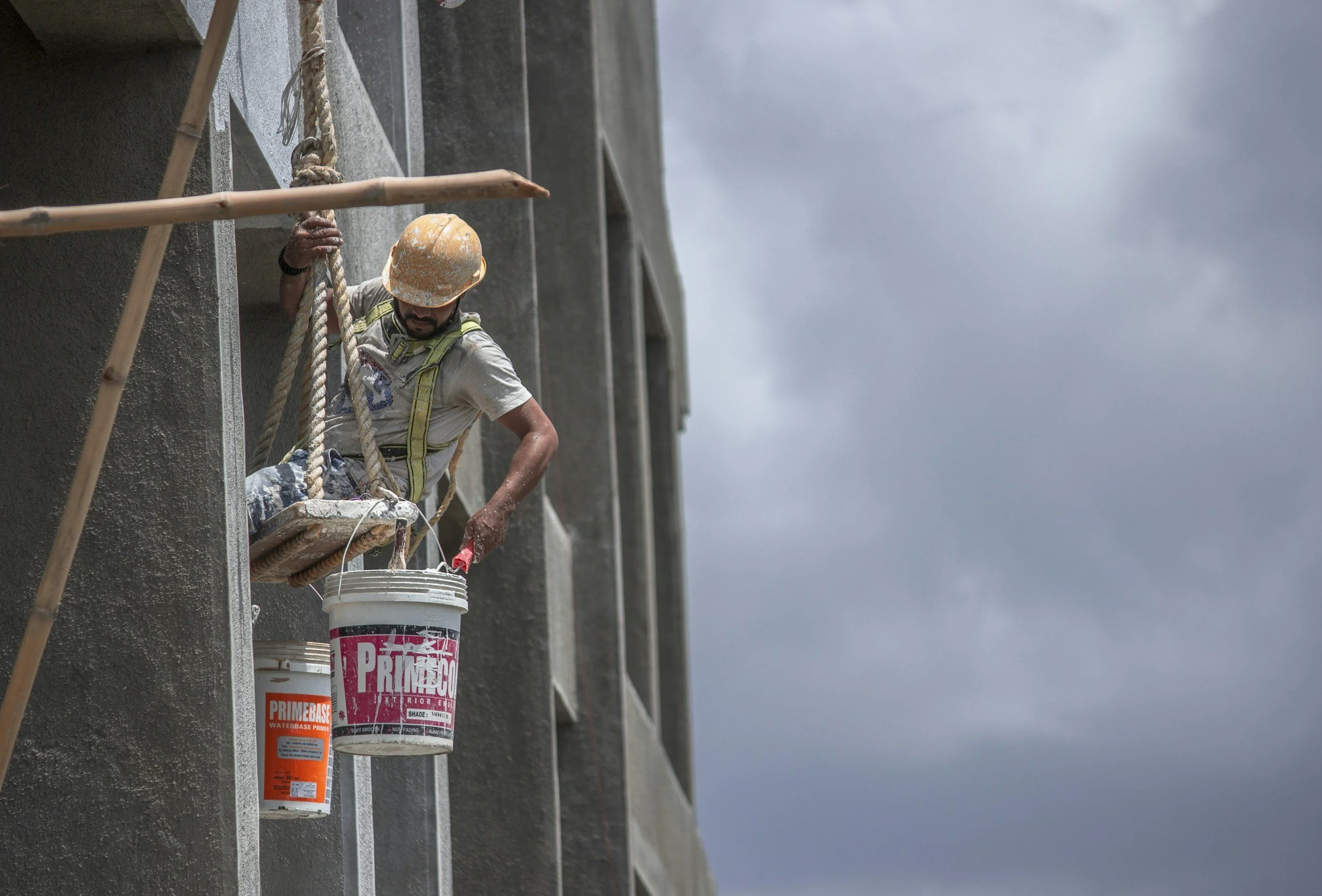 A construction worker with a yellow hard hat and safety harness, sitting on a suspended platform, dipping a paintbrush into a bucket of primer on a high-rise building under cloudy sky.