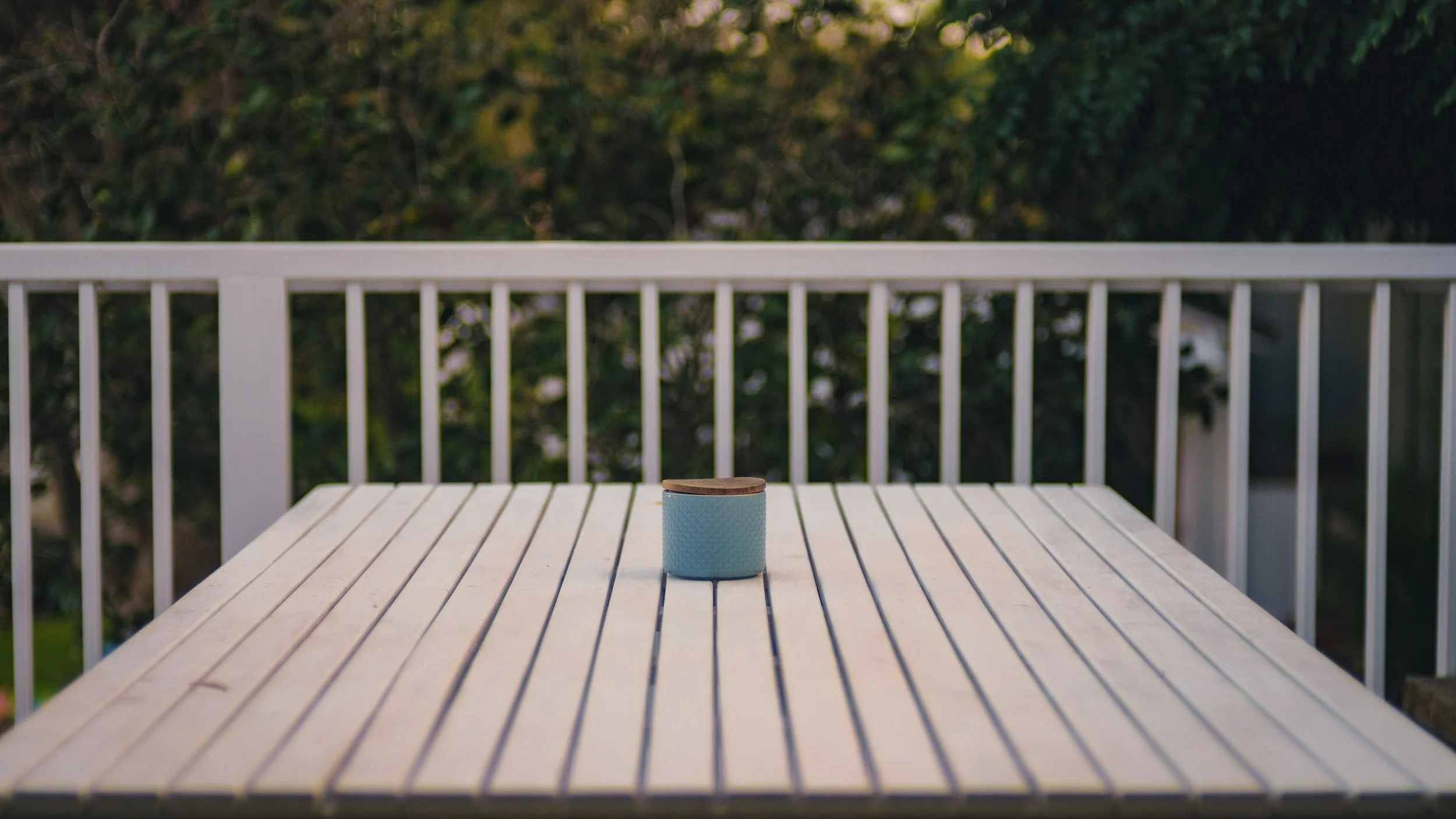 Empty white outdoor table with a small blue container with a wooden lid, on a balcony with white railing, and trees in the background.