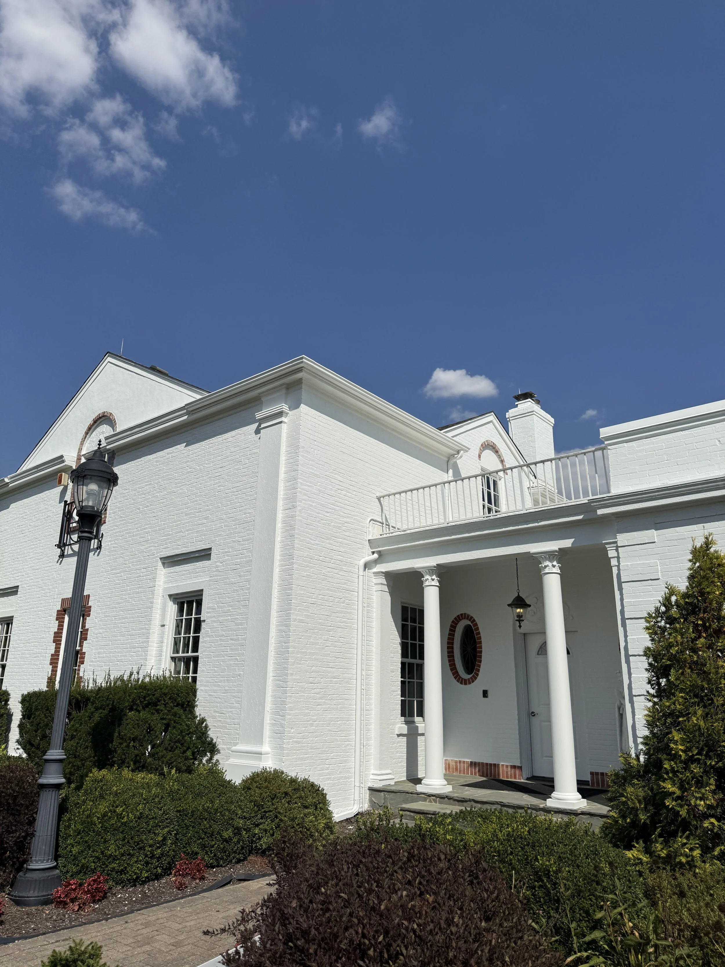 White brick house with columns at the entrance, surrounded by bushes under a blue sky with scattered clouds.