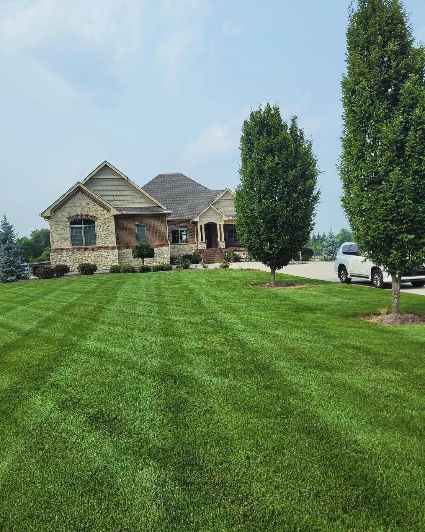 Front yard of a house with a well-maintained green lawn, two trees, a house with a brick and stone facade, a driveway with a parked white vehicle, and a cloudy sky.