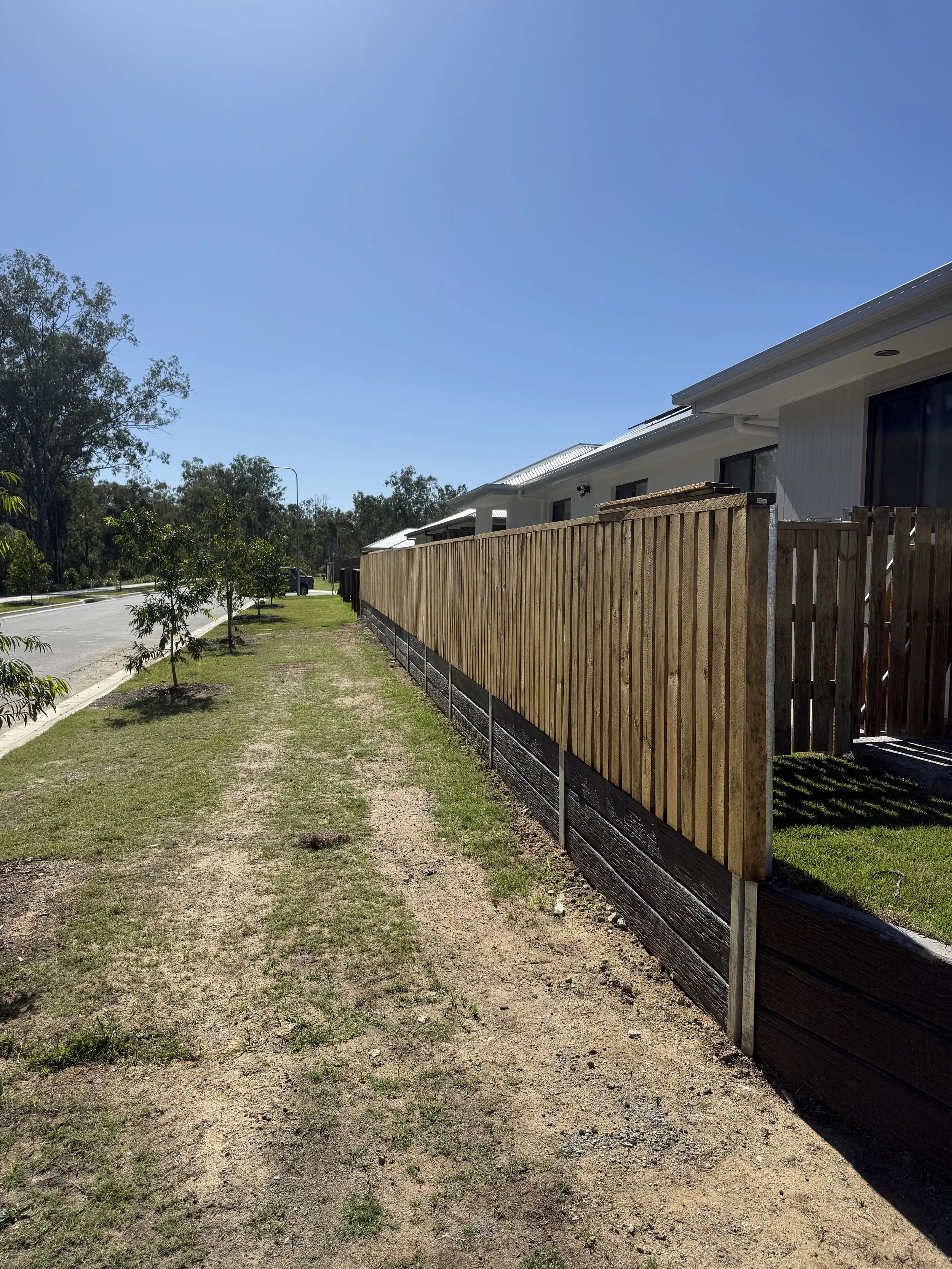 A sidewalk with sparse grass and patches of dirt, lined with small trees on the left, and a new wooden fence on the right separating a house from the sidewalk, under a clear blue sky.