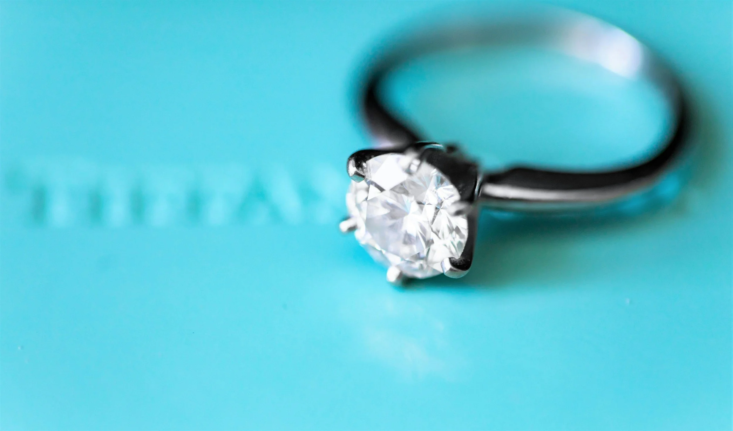 Close-up of a silver Tiffany's engagement ring with a large, round, clear diamond in a prong setting, set against a light blue background. It is the classic Tiffany setting with 6 prongs over the classic Tiffany blue engagement ring box