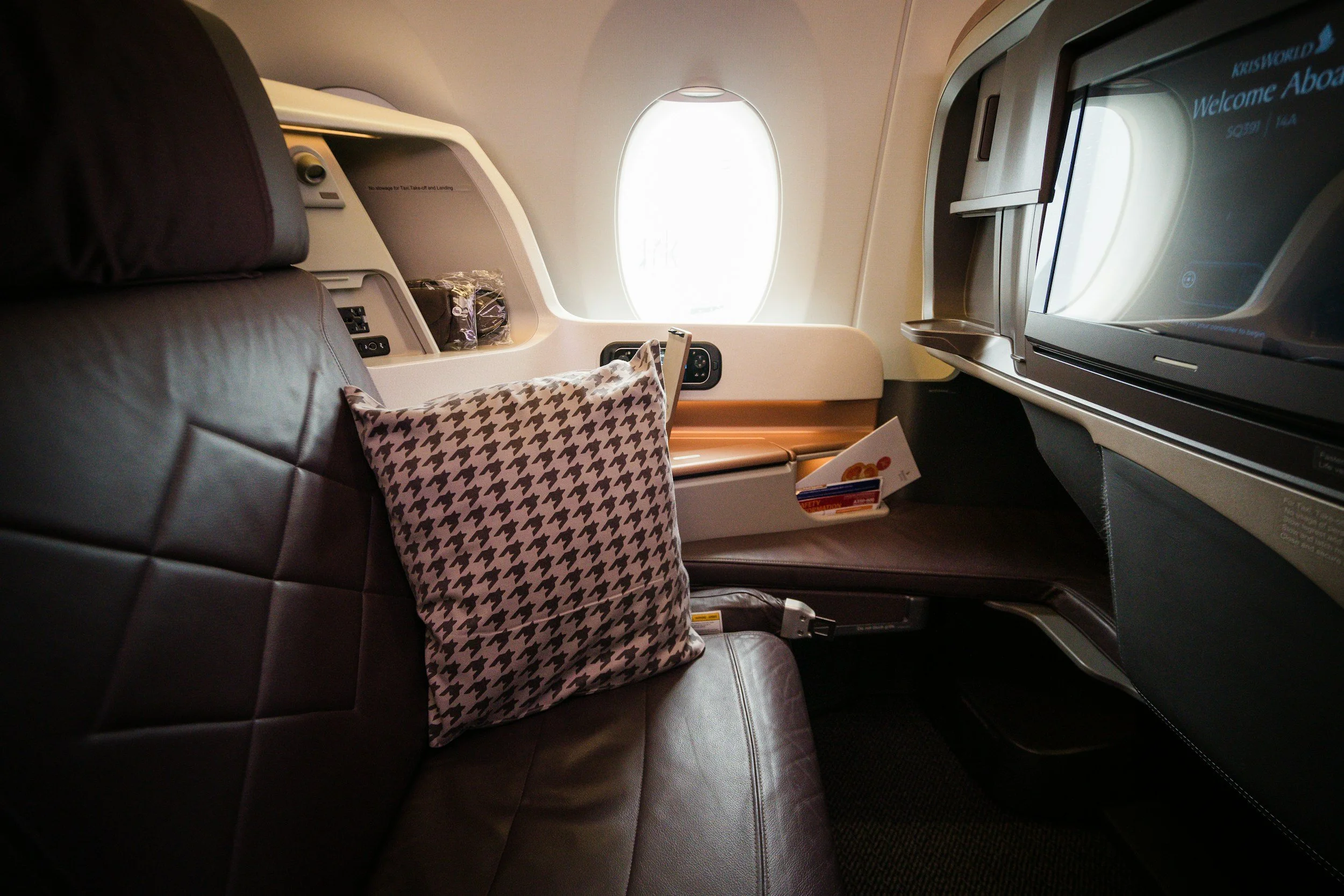 First class airplane seat with a pillow, window, and in-flight entertainment screen.