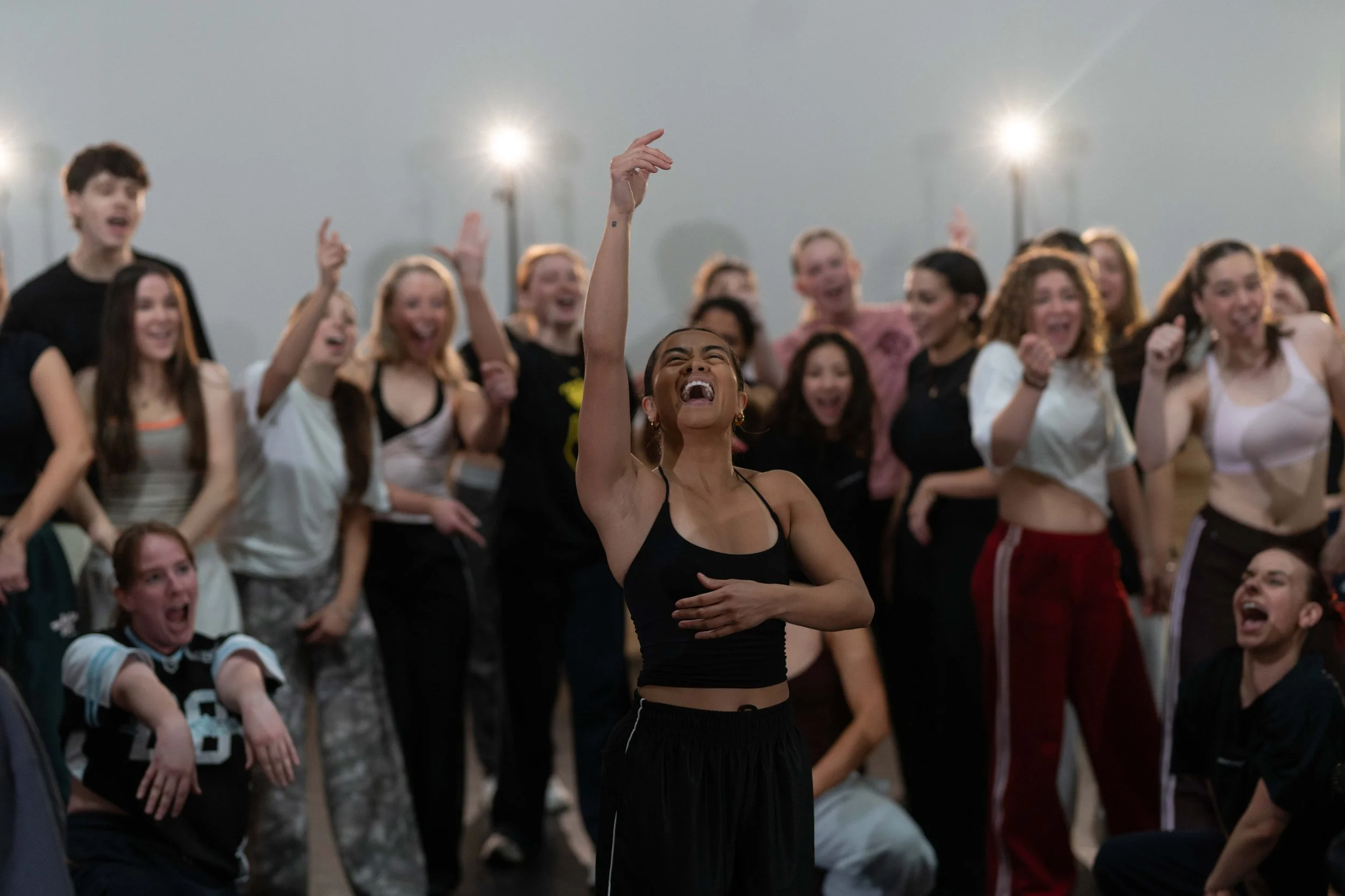 A group of people smiling and laughing, with a woman in the front raising her hand and holding her stomach, in a large indoor space with bright lights.