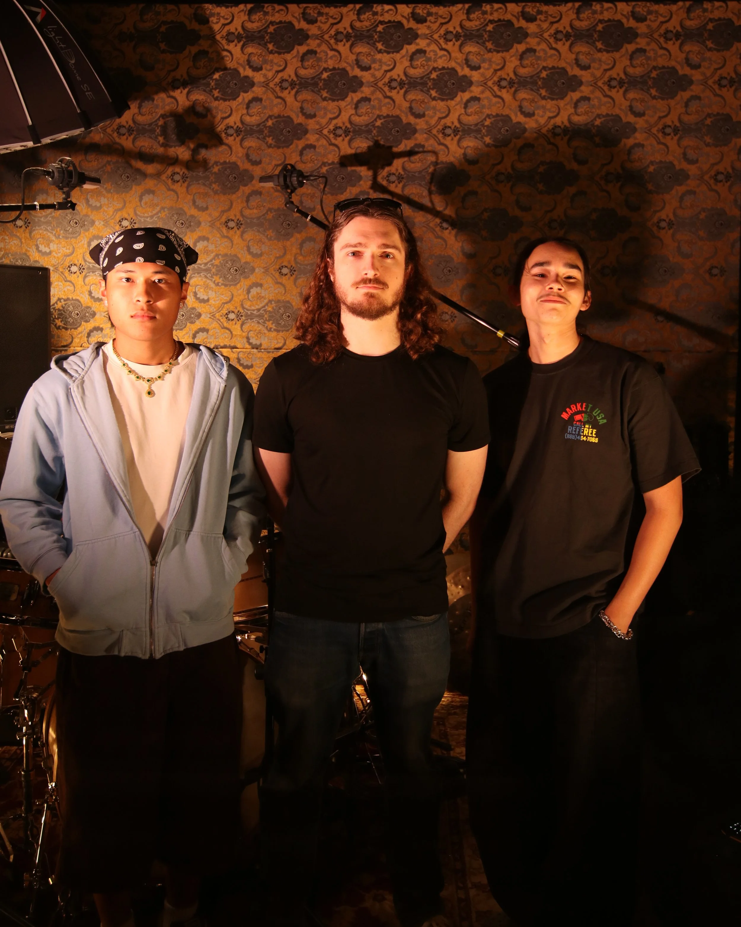 Three young men standing side by side in front of a patterned wall. The man on the left wears a black bandana, white jacket, and a necklace. The man in the middle has long curly hair, a beard, sunglasses on his head, and a plain black t-shirt. The man on the right has short hair, a mustache, a black t-shirt with colorful text, and a chain bracelet on his right wrist. Studio lighting and equipment are visible behind them.