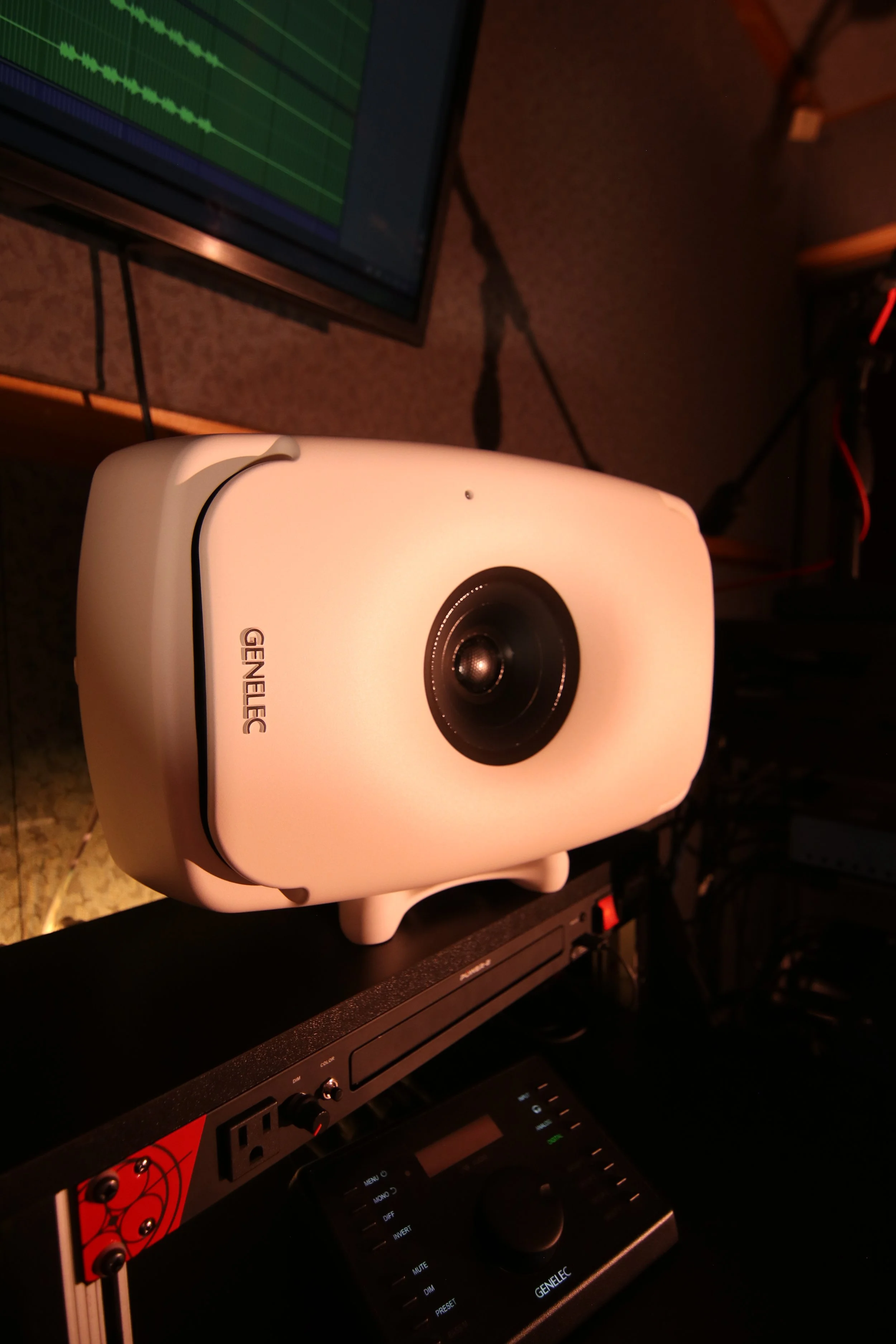 A Genelec monitor speaker facing an electronic music mixing controller, with a computer screen displaying waveform audio editing in the background.