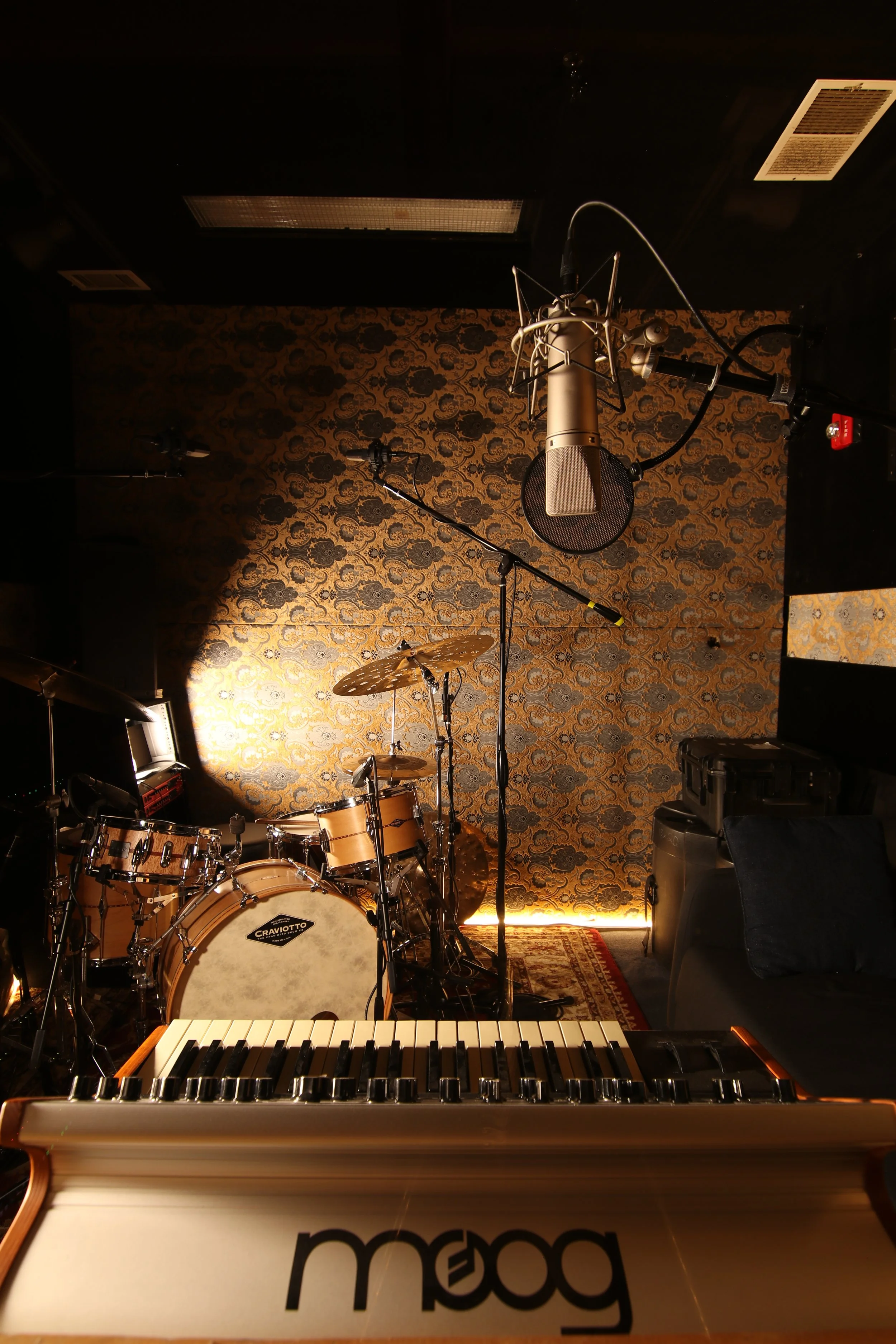 A music studio with a drum set, keyboard, microphone, and various audio equipment. The wall has a decorative pattern, and there are stage lights illuminating part of the room.