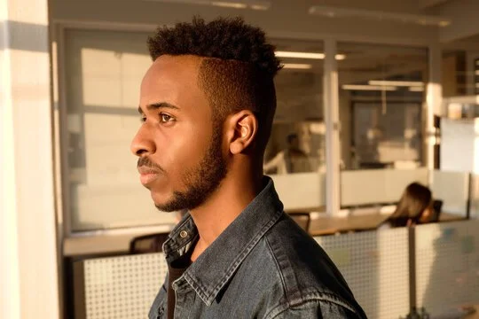A young man with a fade haircut and a beard looking to the left, standing in an indoor space with large windows.