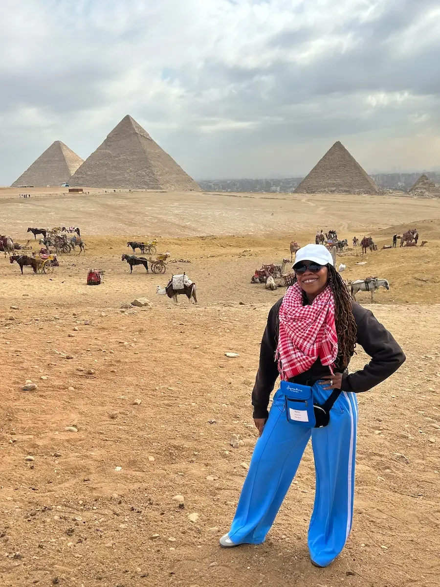 Person stands smiling in a desert with pyramids in the background, conveying confidence and a sense of exploration.