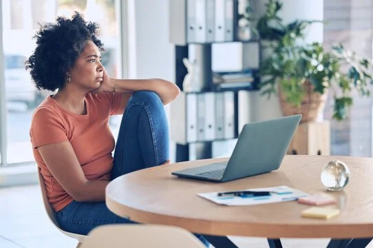A woman sitting at a table with a laptop, looking thoughtful or pensive.