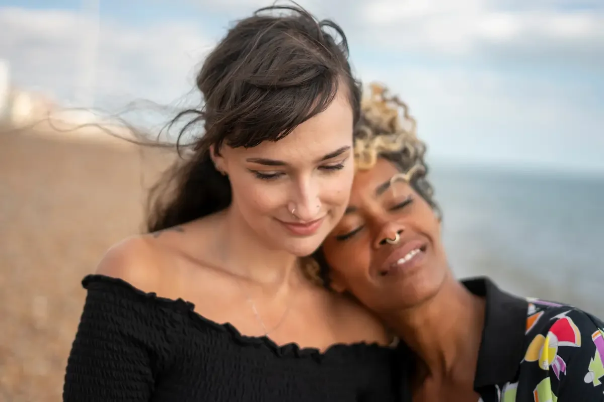 Two women with nose piercings and closed eyes smiling and leaning into each other on a beach with ocean in the background.