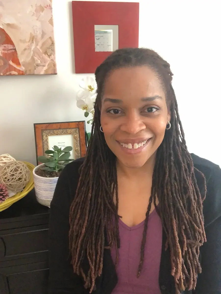 Raven Waterman smiles warmly at the camera, seated indoors beside plants and framed art in a calm, welcoming space.