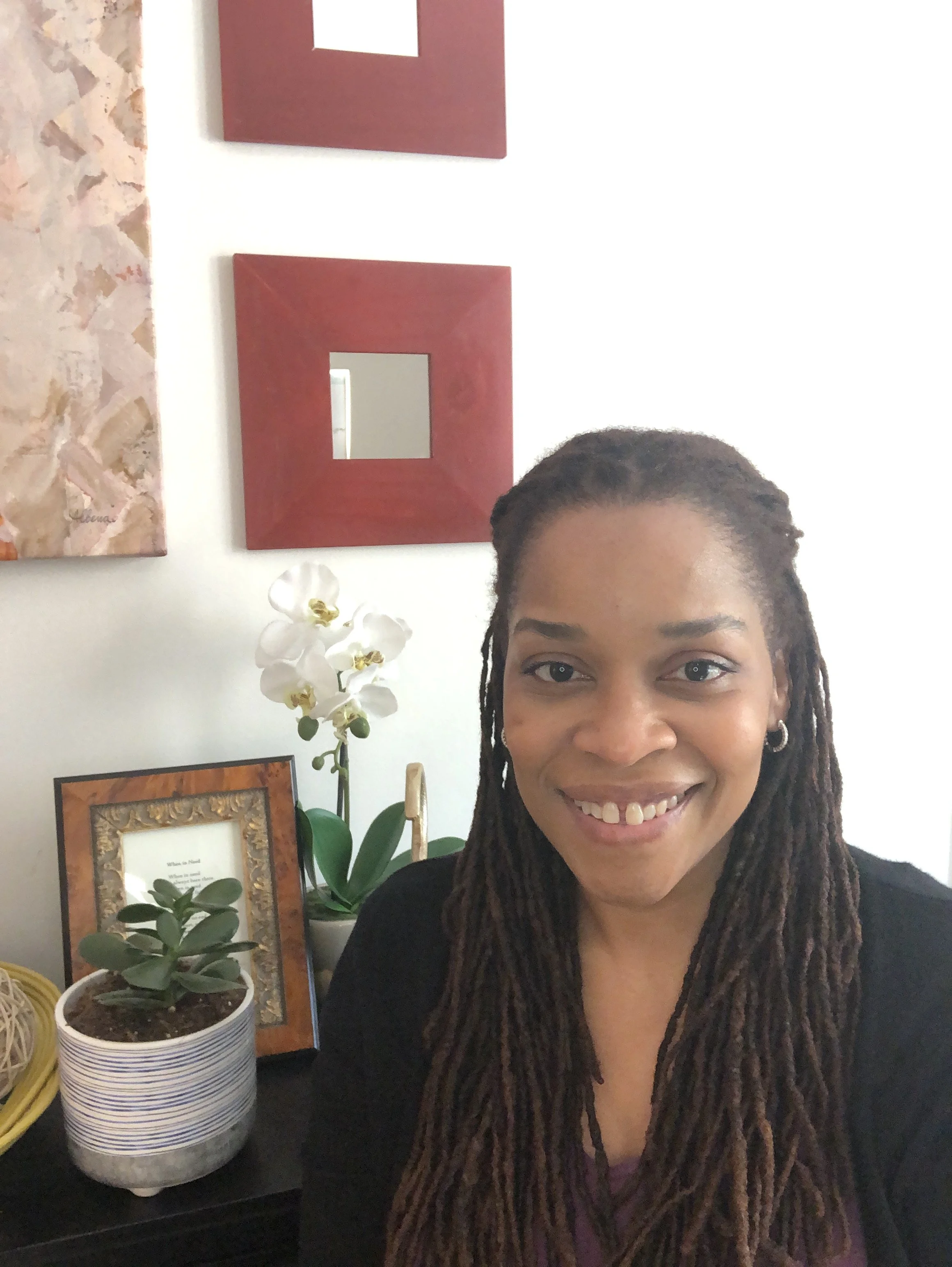 Woman with long dreadlocks smiling in a room decorated with framed artwork, plants, and orchids.