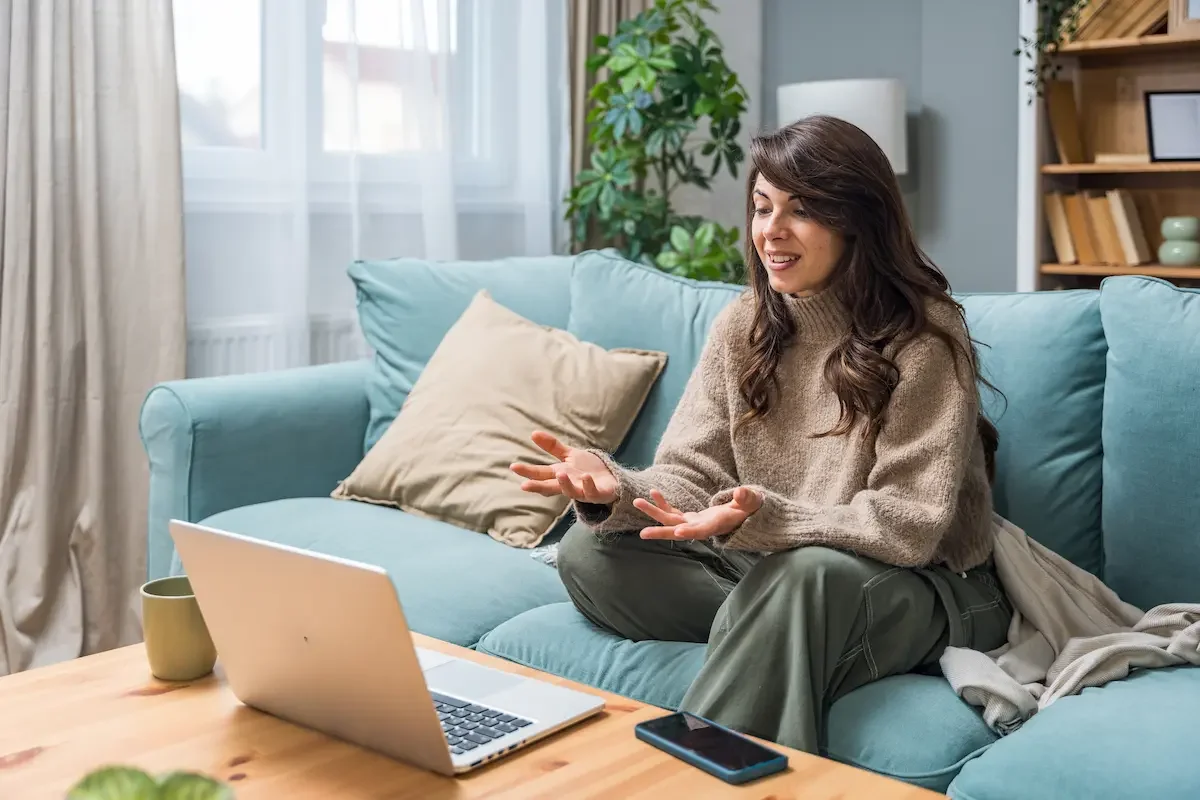 A woman sitting on a light blue sofa, smiling and gesturing with her hands, in a living room with a laptop, a coffee mug, and a smartphone on the table in front of her.
