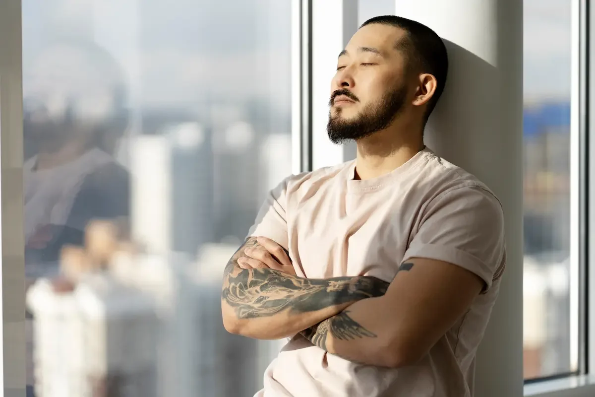 A man with tattoos and a beard standing with arms crossed, eyes closed, leaning against a window with a cityscape in the background, enjoying sunlight.