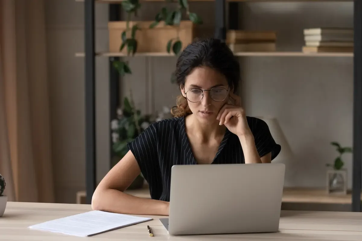 A woman with glasses and dark hair sits at a desk, working on a laptop, with papers and a pen in front of her, in a room with shelves and plants in the background.