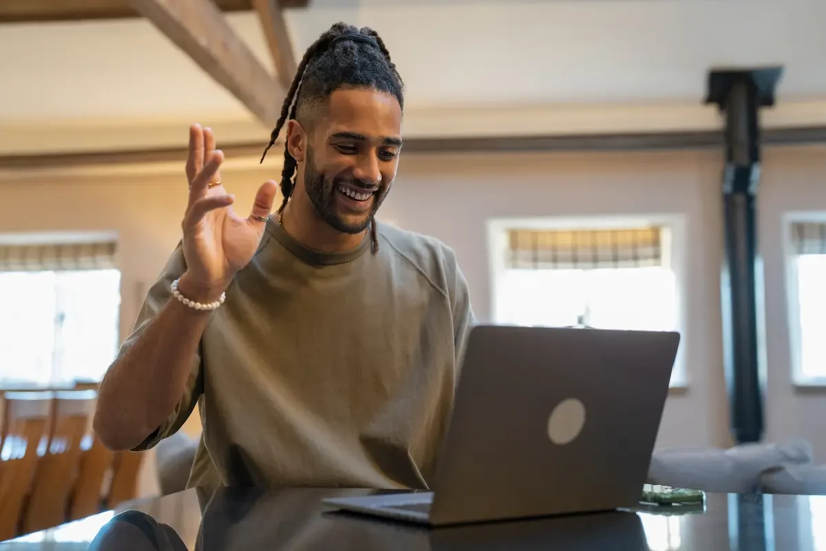 A young man with dreadlocks smiling and waving at his laptop in a bright room.