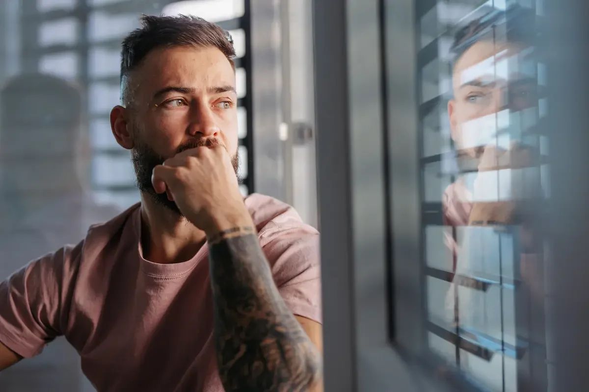 A man with a beard and tattoos on his arm is looking thoughtfully out a window in a modern building, with his hand resting on his chin.