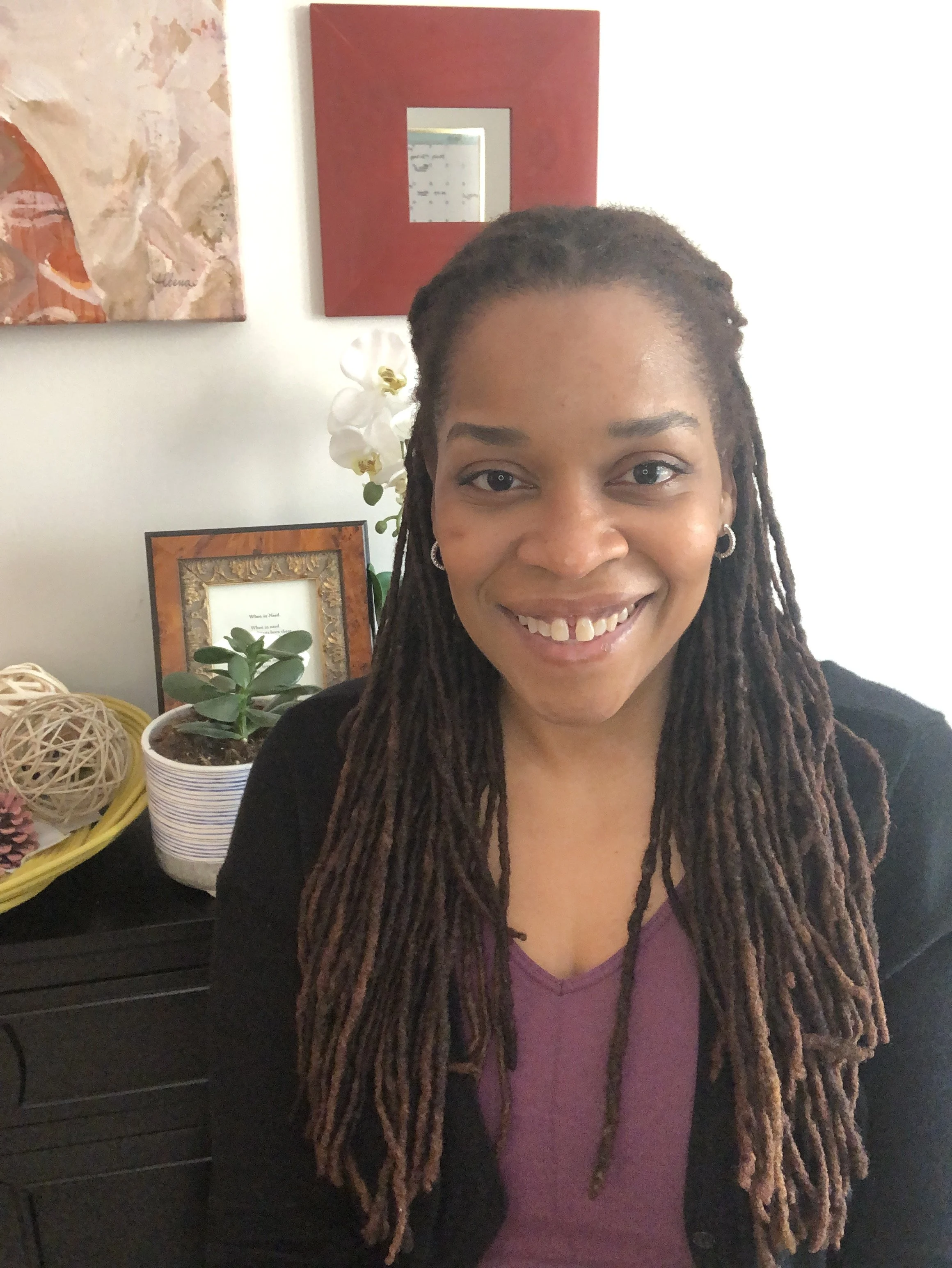 A woman with long dreadlocks, wearing a purple top and black jacket, smiling at the camera. Behind her, there are various decorative items including a framed picture with green plants, a white orchid, a yellow basket with decorative balls, and abstract wall art.