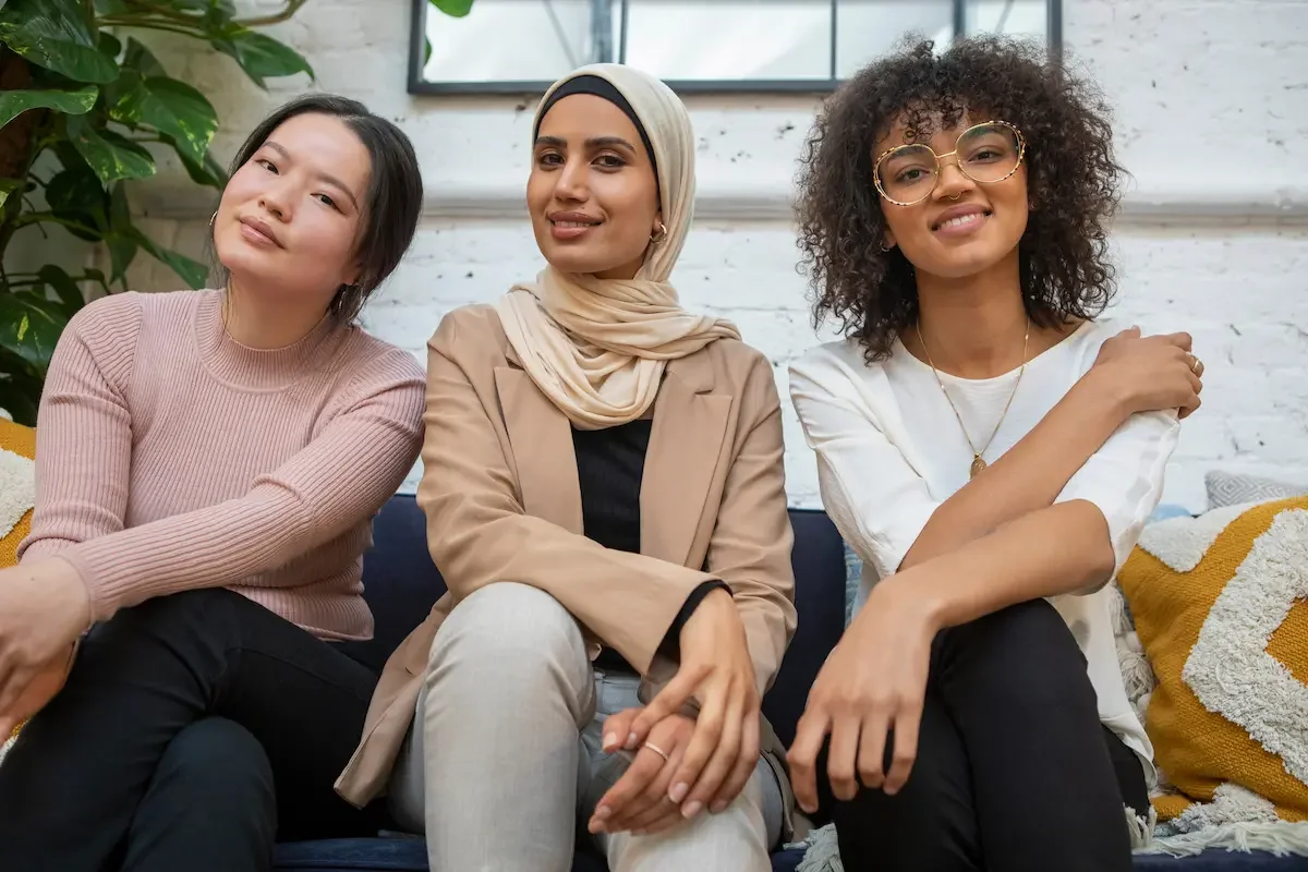Three women sitting close together on a couch, smiling at the camera, in a bright room with white brick walls and a green plant in the background.
