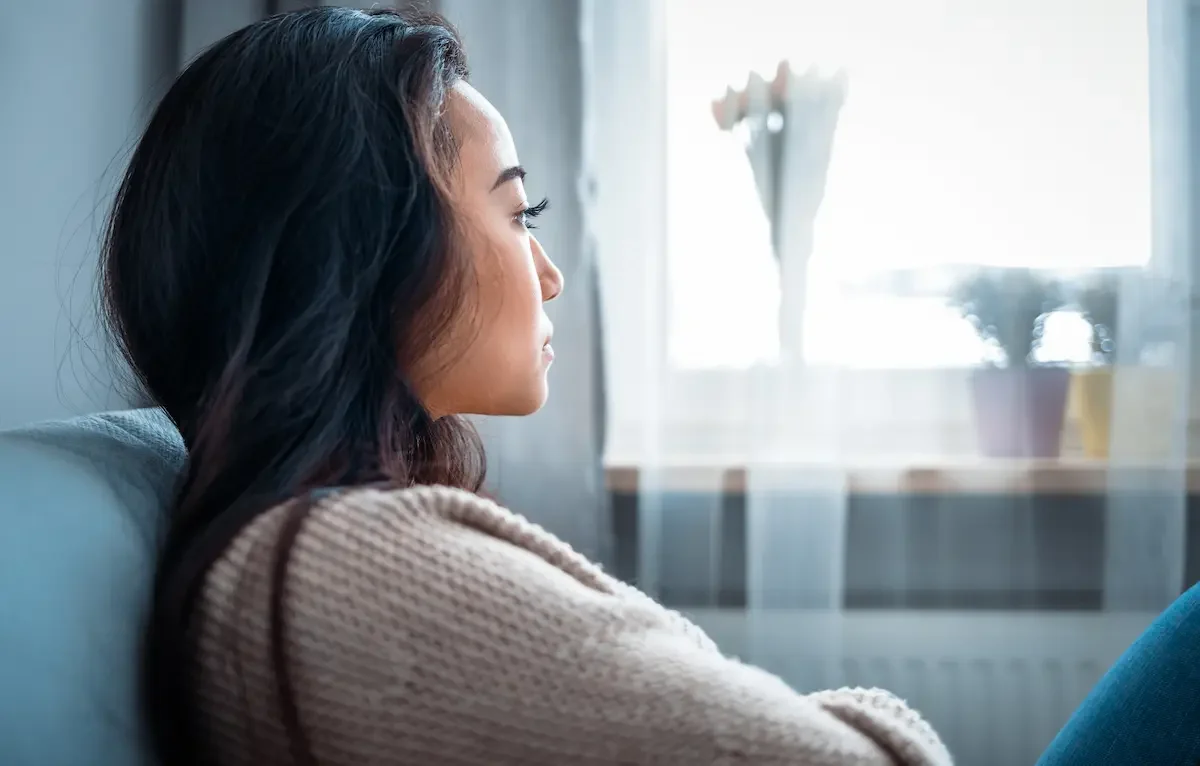 A woman with long dark hair sitting on a couch, looking thoughtfully out of a window with sheer curtains, in a cozy room with plants on the windowsill.