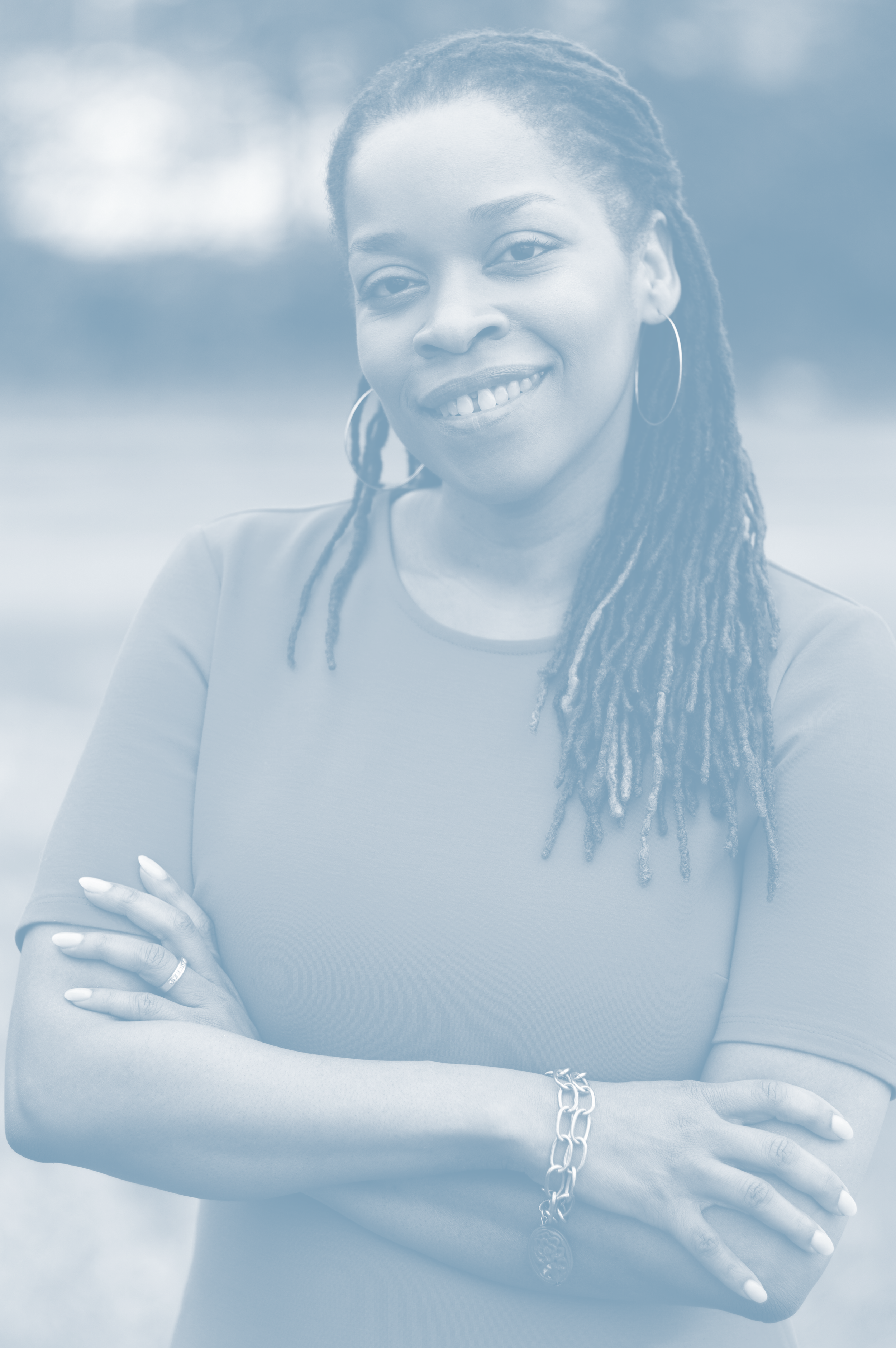 Portrait of a woman with long dreadlocks, wearing hoop earrings, a bracelet, and a short-sleeve shirt, standing outdoors with a blurred background.