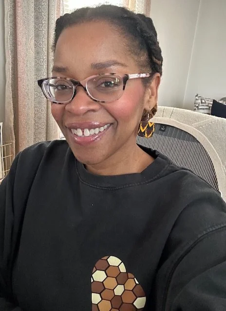 A woman with glasses and earrings smiling at the camera in a room with curtains and a chair.