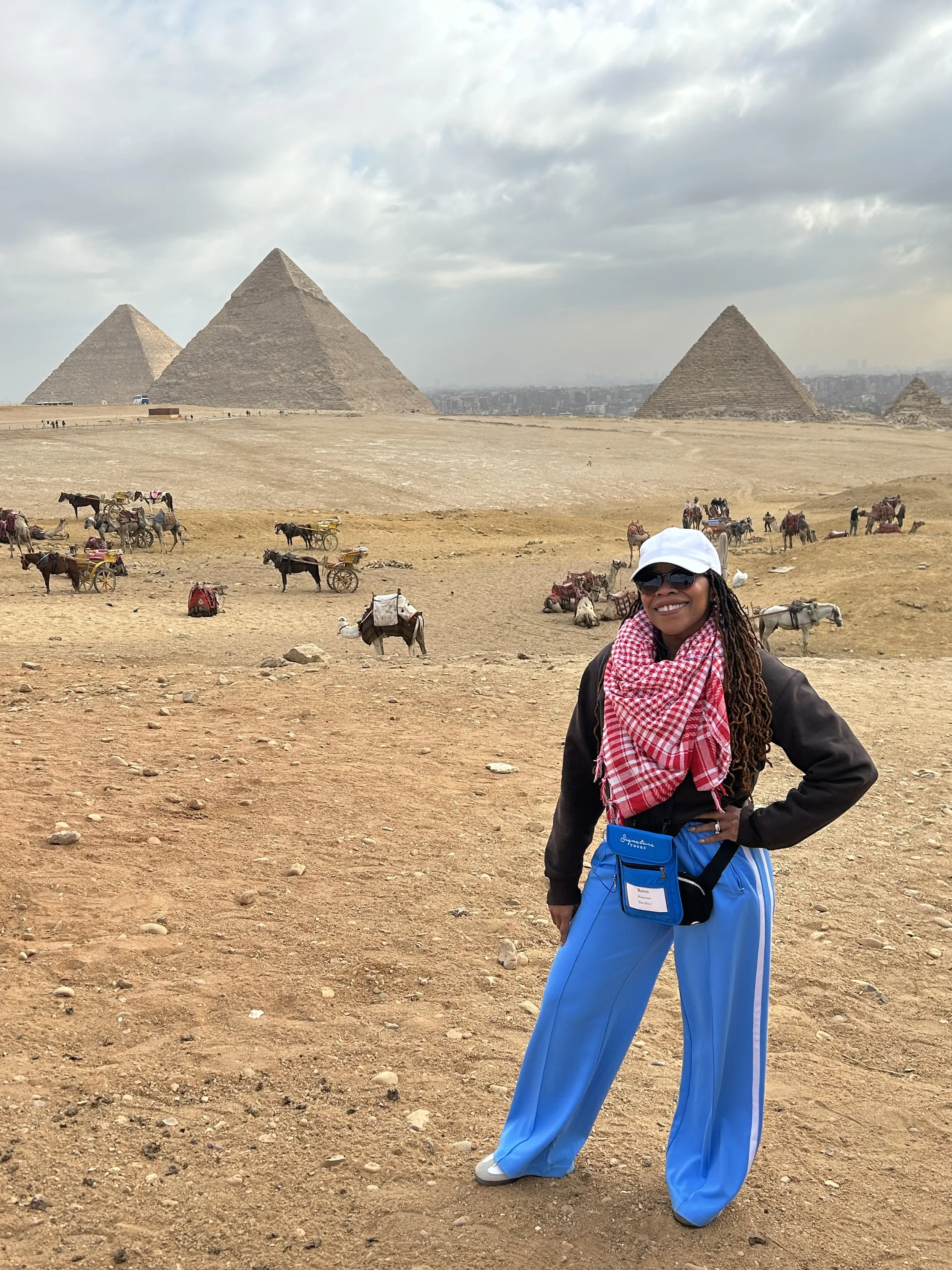 A woman smiling and posing in front of the Pyramids of Giza in Egypt, wearing a white cap, sunglasses, a red and white scarf, a black jacket, and blue pants, with camels and tourists in the background under a cloudy sky.