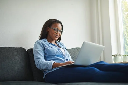 A woman with glasses, wearing a denim shirt, sitting on a dark couch, working on a laptop in a bright room with a large window and potted plants.