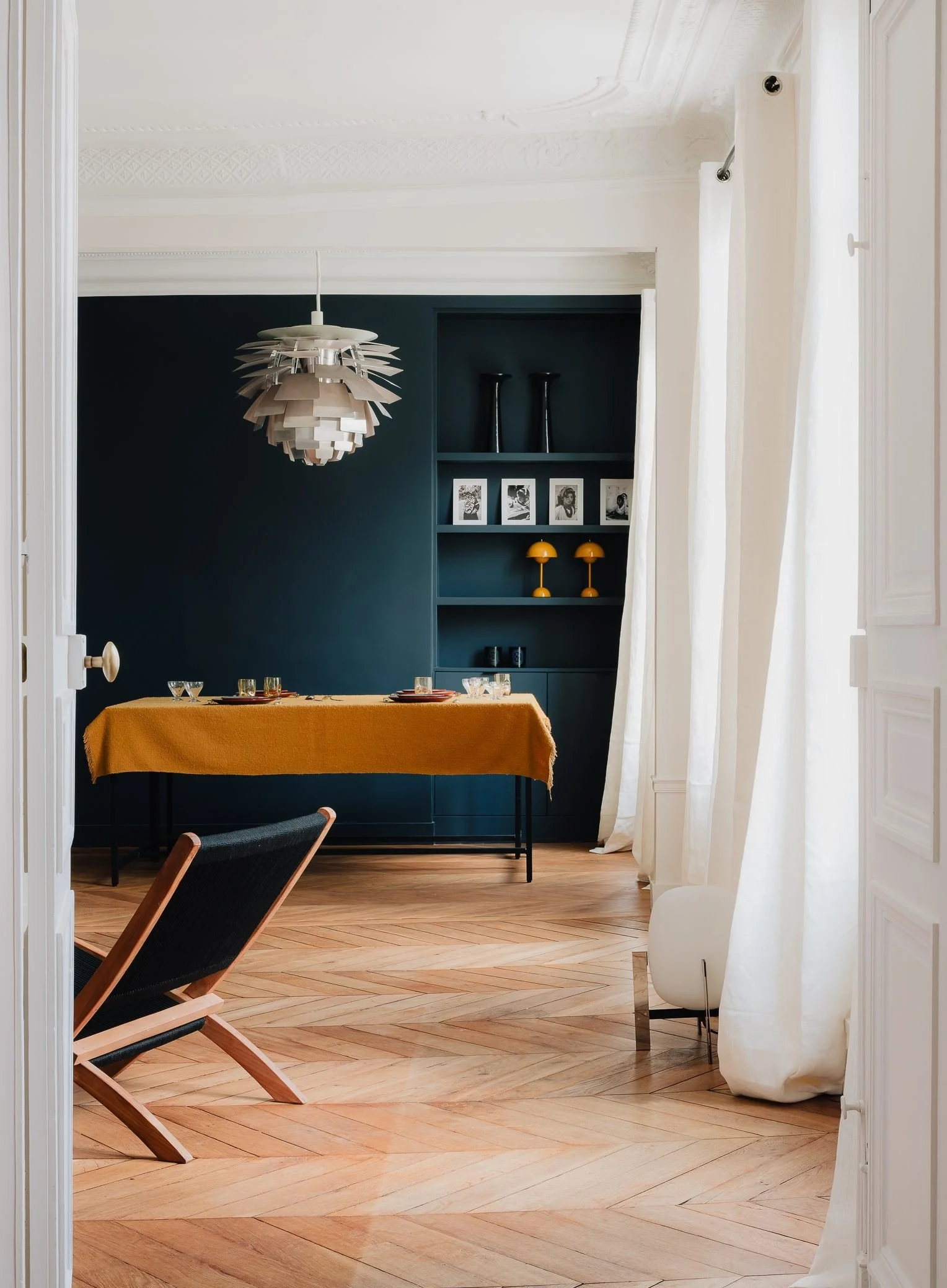 Architecte d’intérieur à Paris, salle à manger  à manger avec mur bleu foncé, Louis poulsen PH Artichoke Luminaires, chaises en bois et tissu noir, étagère avec photos en noir et blanc, lampadaire blanc et suspensions jaunes, rideaux blancs, parquet