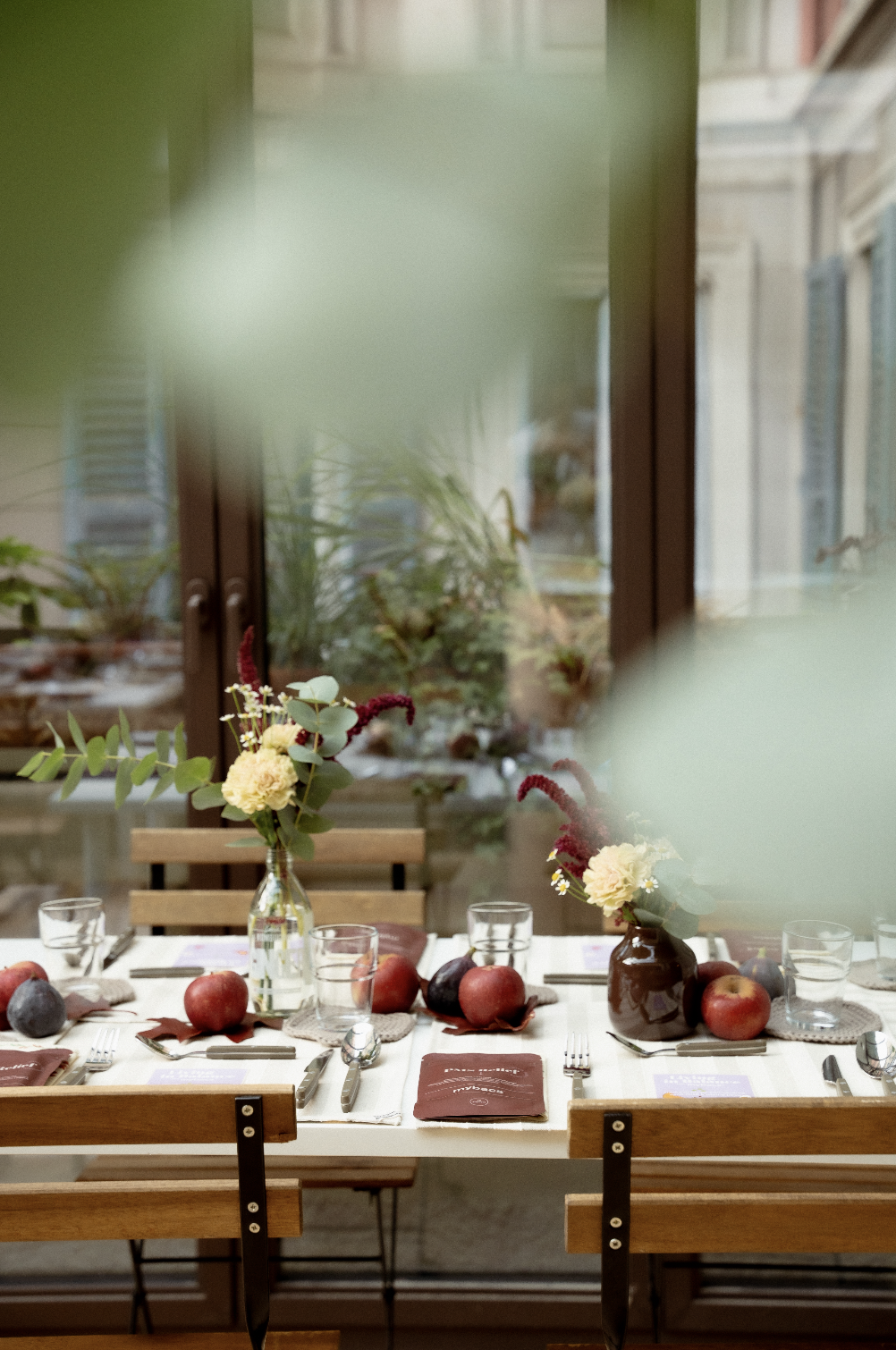 A restaurant table decorated with floral centerpieces, apples, glasses, and cutlery, placed near a window with green foliage outside.