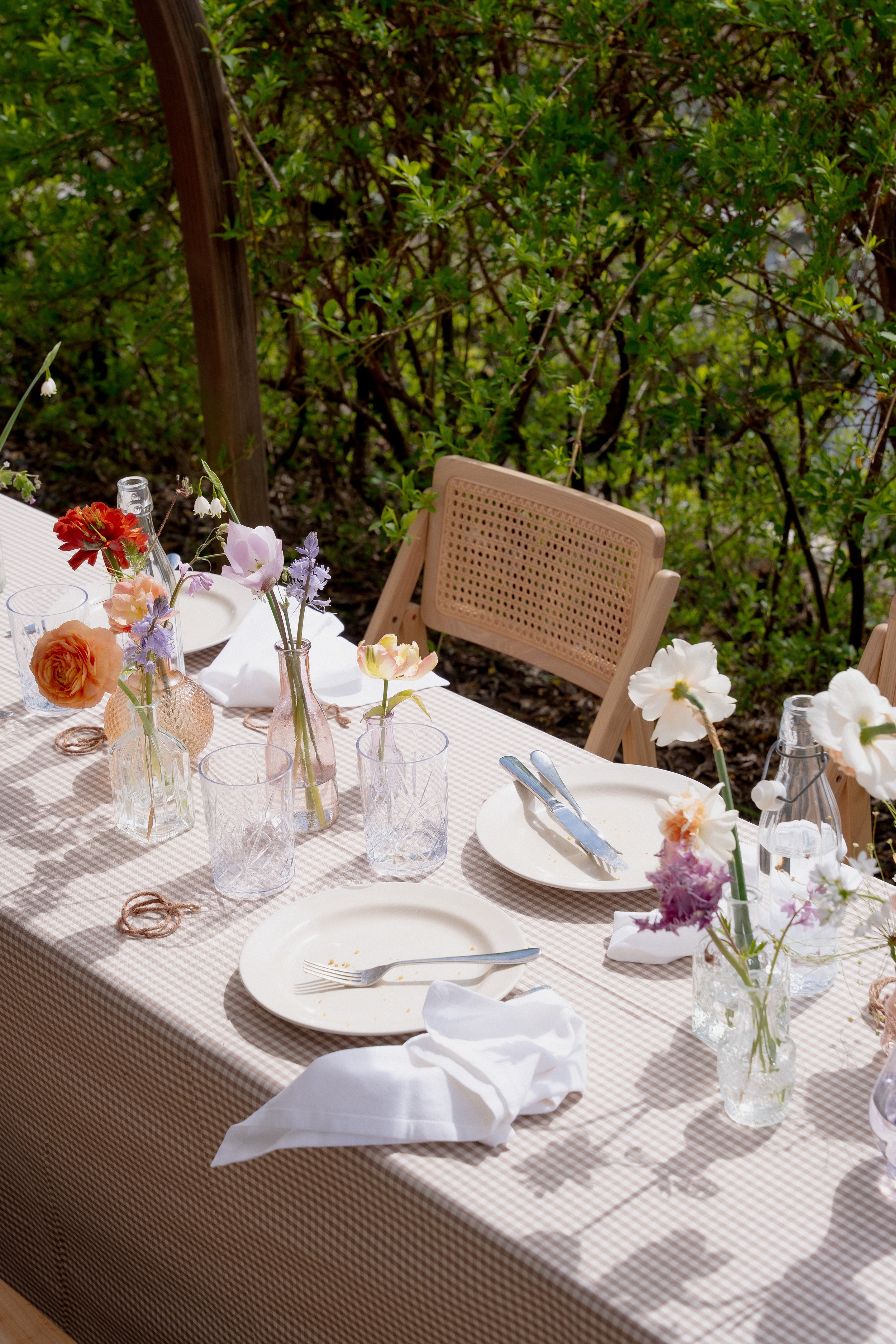 Outdoor dining table set with white plates, utensils, napkins, and a variety of colorful flowers in glass vases on a checkered tablecloth, surrounded by wooden chairs and greenery.