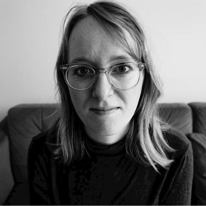 A black and white image of a woman in glasses sat on a sofa.
