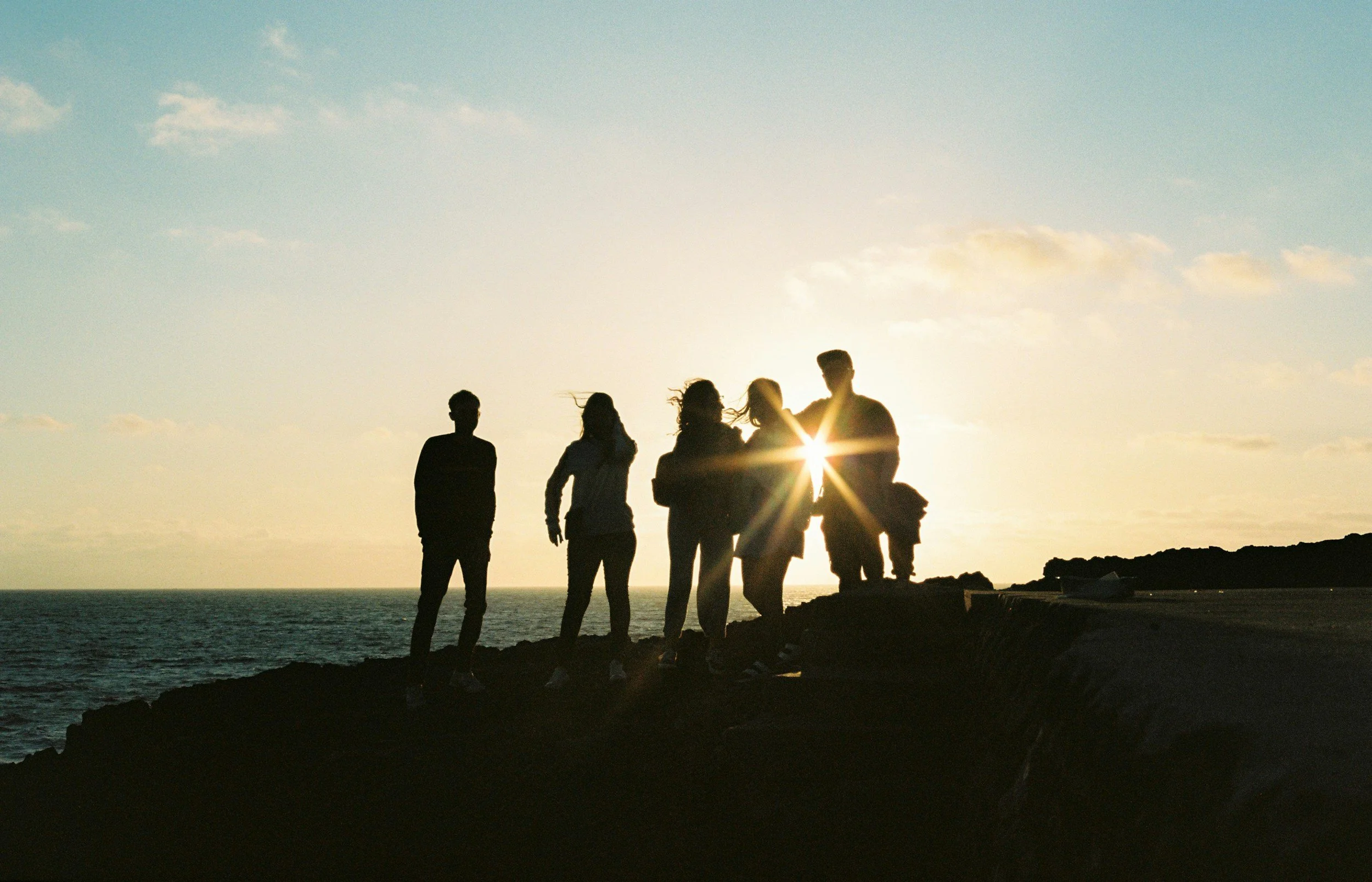 Group of people standing on a rocky shoreline at sunset, silhouetted against the sky with the sun shining through gap between two individuals.
