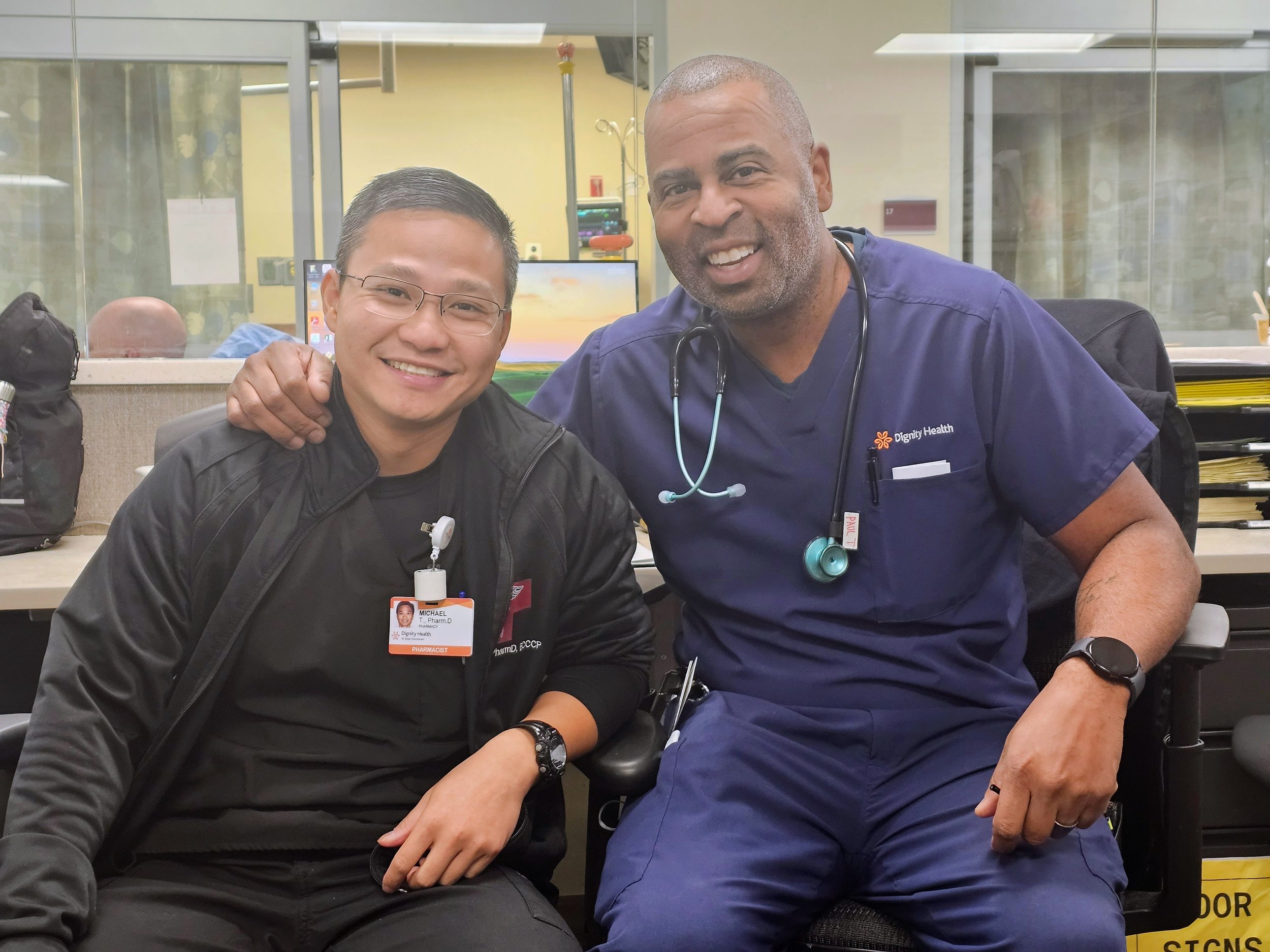 Two smiling healthcare professionals, one wearing a black jacket with a name badge and the other in blue scrubs with a stethoscope, sitting in a medical office or hospital setting.