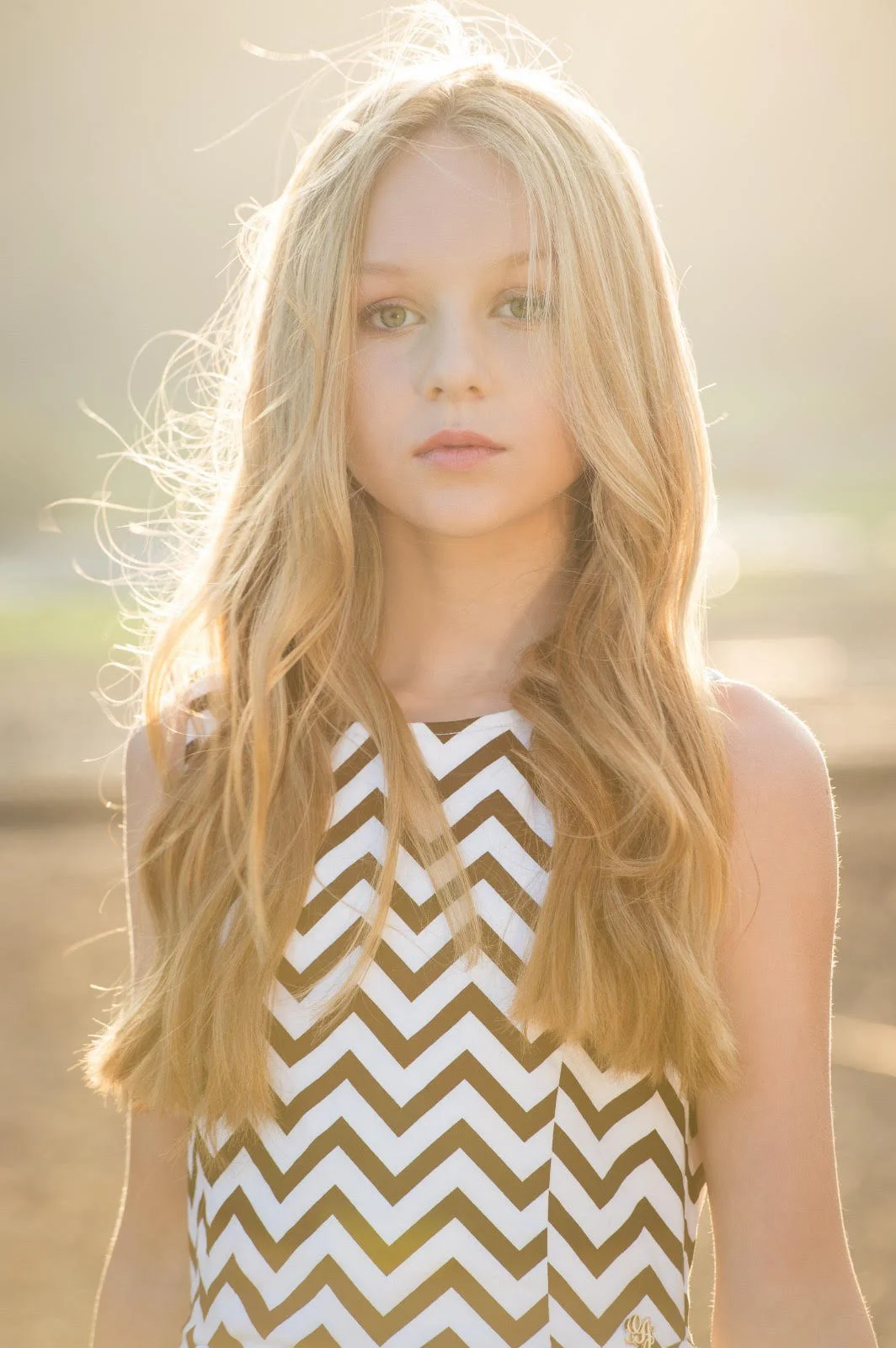 A young woman with long blonde hair and green eyes standing outdoors in sunlight, wearing a sleeveless black and white zigzag patterned top.