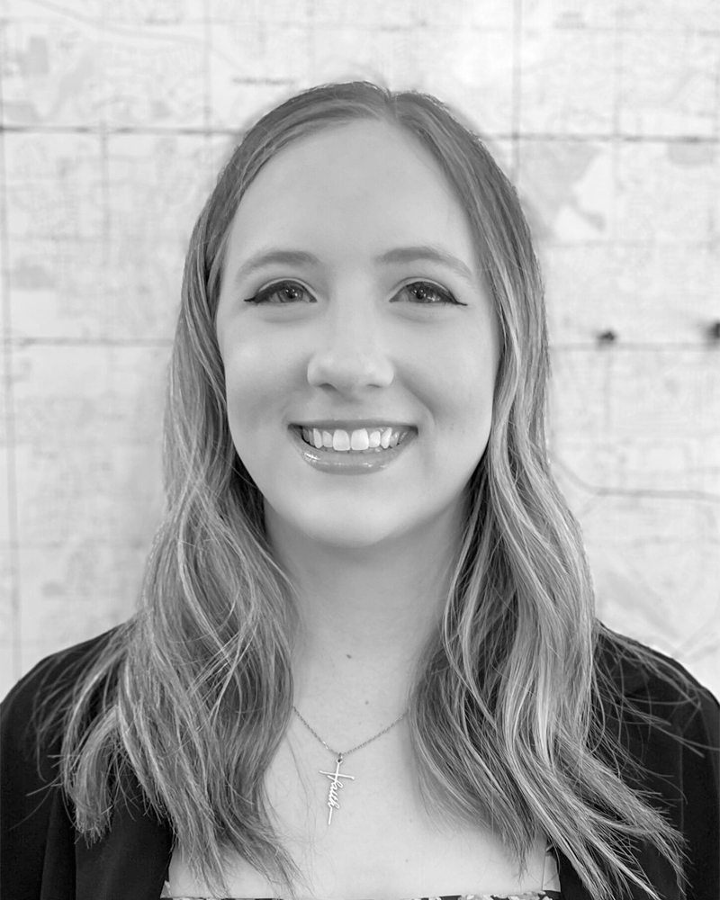 Black and white portrait of a smiling woman with wavy hair, wearing a necklace with a cross pendant, standing in front of a wooden wall.