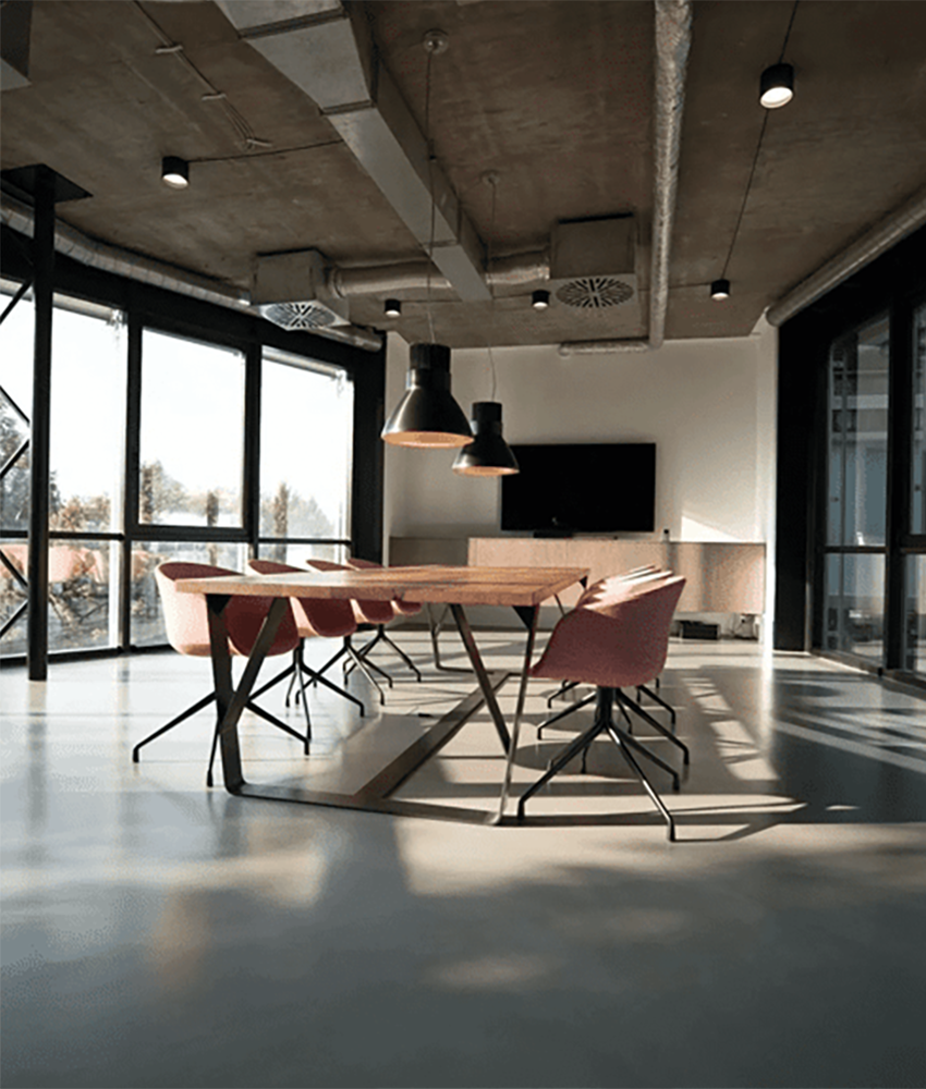 Modern conference room with a large wooden table, pink chairs, pendant lighting, and a wall-mounted TV, with large windows letting in natural light.