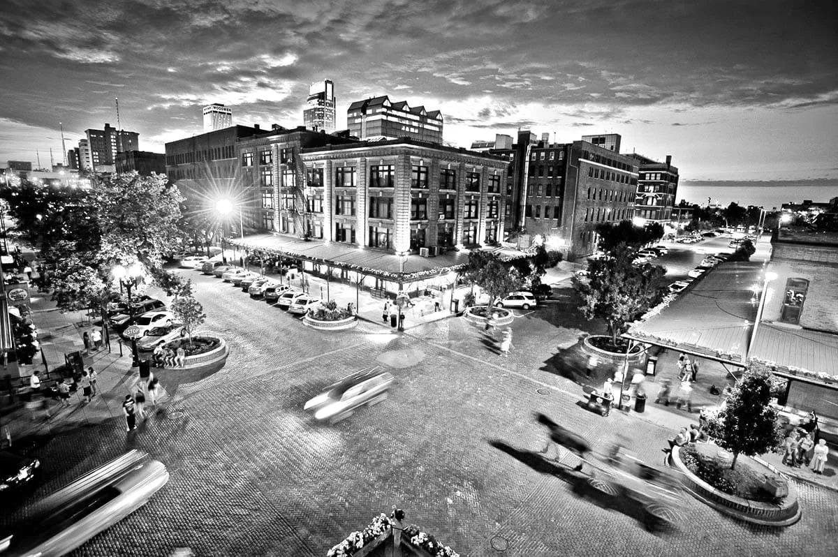 Black and white photo of an urban city scene at dusk, with busy streets, parked cars, pedestrians, trees, and multi-story buildings with lit windows.
