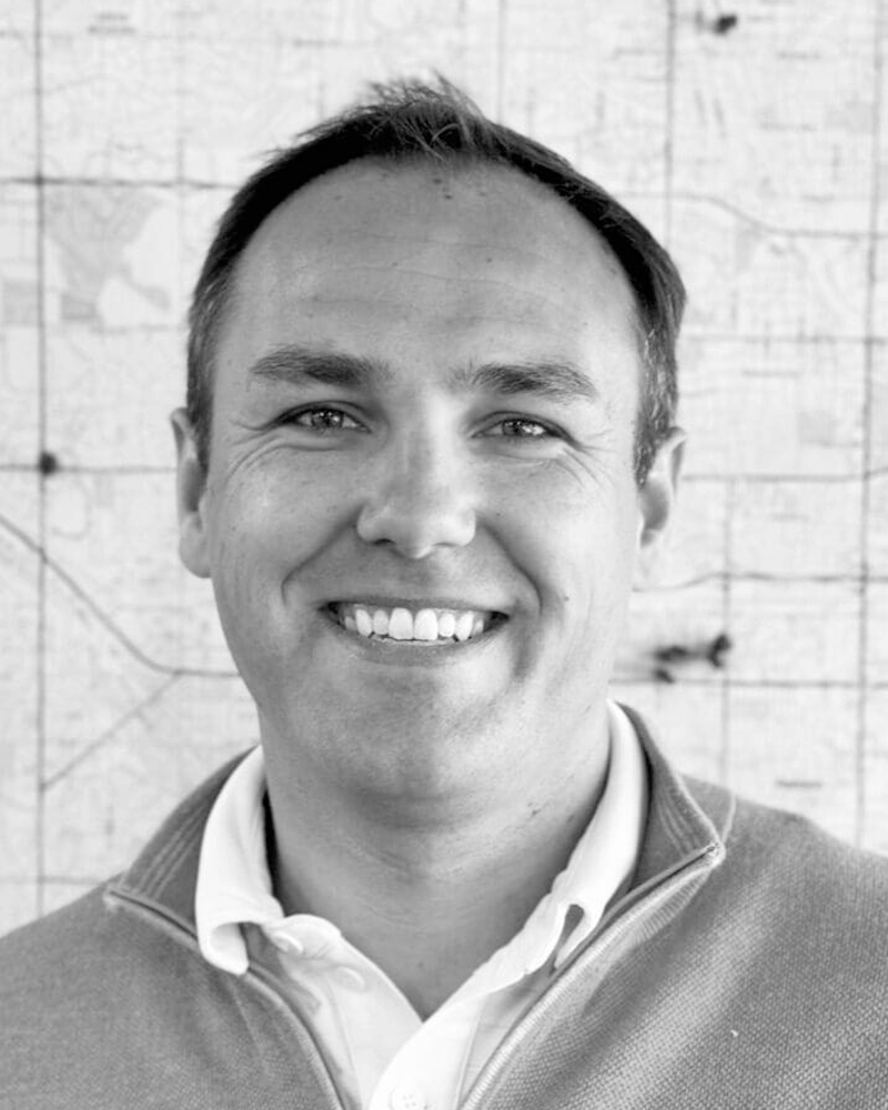 Black and white portrait of a smiling man in casual clothing, standing in front of a textured wall.
