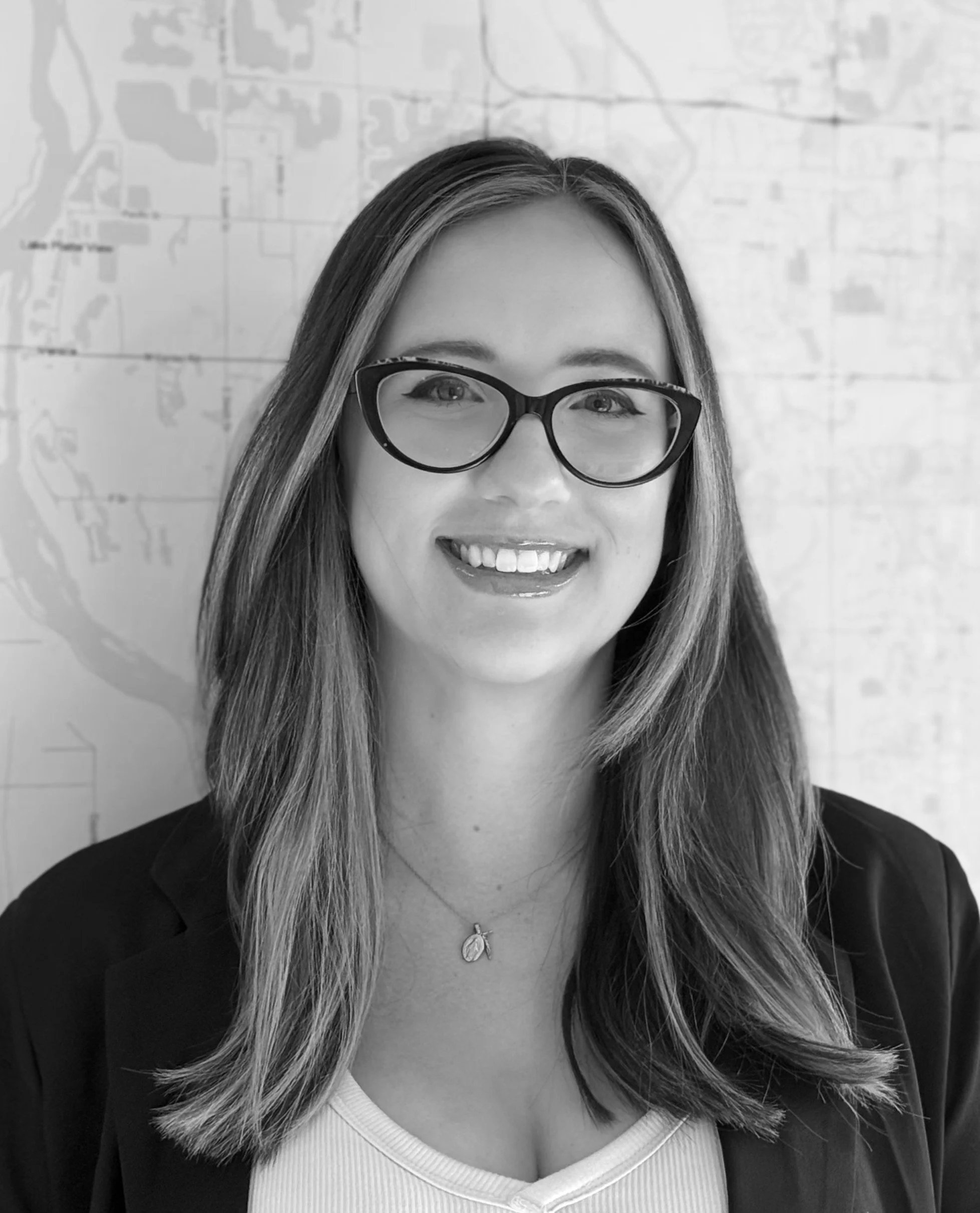 Black and white portrait of a smiling woman with wavy hair, wearing a necklace with a cross pendant, standing in front of a wooden wall.