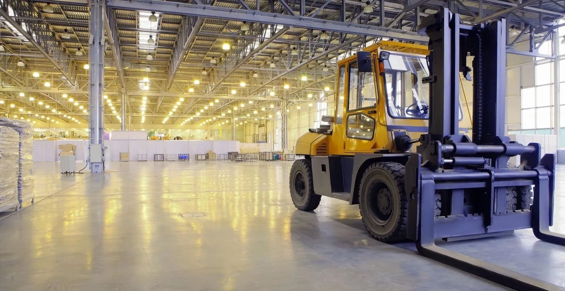 Inside a spacious warehouse with bright overhead lighting, a yellow and black forklift is parked on the shiny concrete floor.