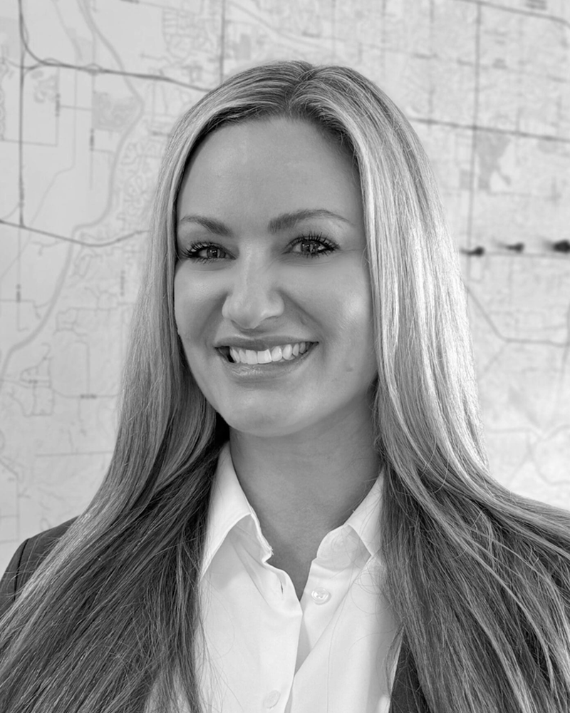 Black and white photo of a smiling woman with long hair, wearing a suit jacket and collared shirt, standing against a map background.