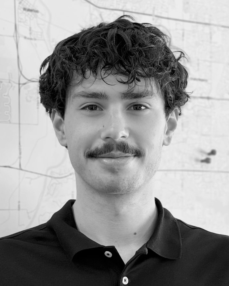 Black and white photo of a young man with curly hair, a mustache, and wearing a collared shirt, standing in front of a map on a wall.