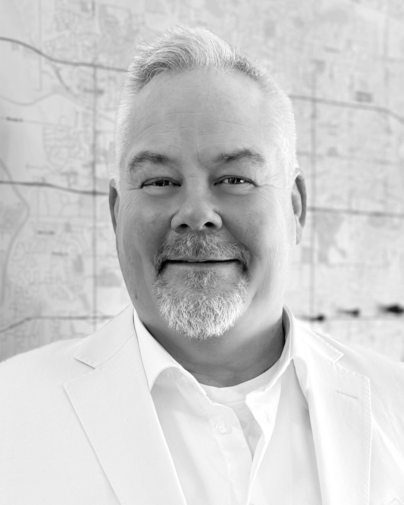 A smiling middle-aged man with gray hair and a beard, wearing a white shirt and blazer, standing in front of a blurred map background.