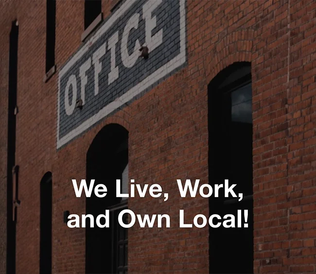 Brick building with an 'OFFICE' sign at the top and text overlay that reads, 'We Live, Work, and Own Local!'
