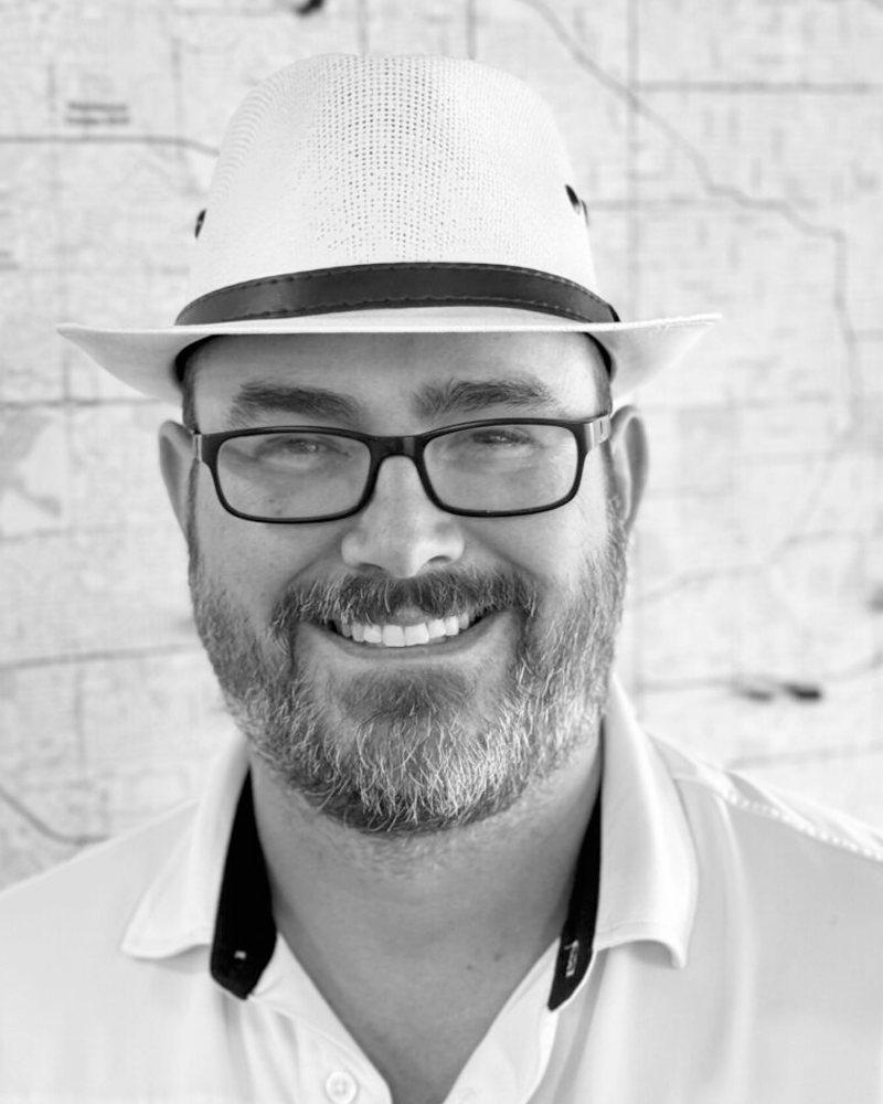 A man with glasses, a beard, and a mustache smiling, wearing a straw hat with a black band, and a collared shirt.