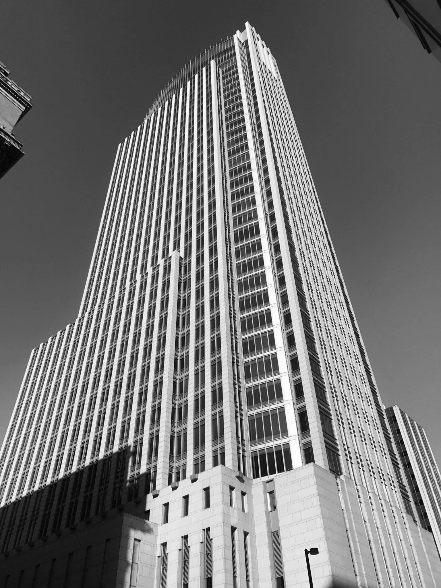 Tall modern high-rise building with glass windows and vertical lines, shadow cast on lower part, clear sky in the background.
